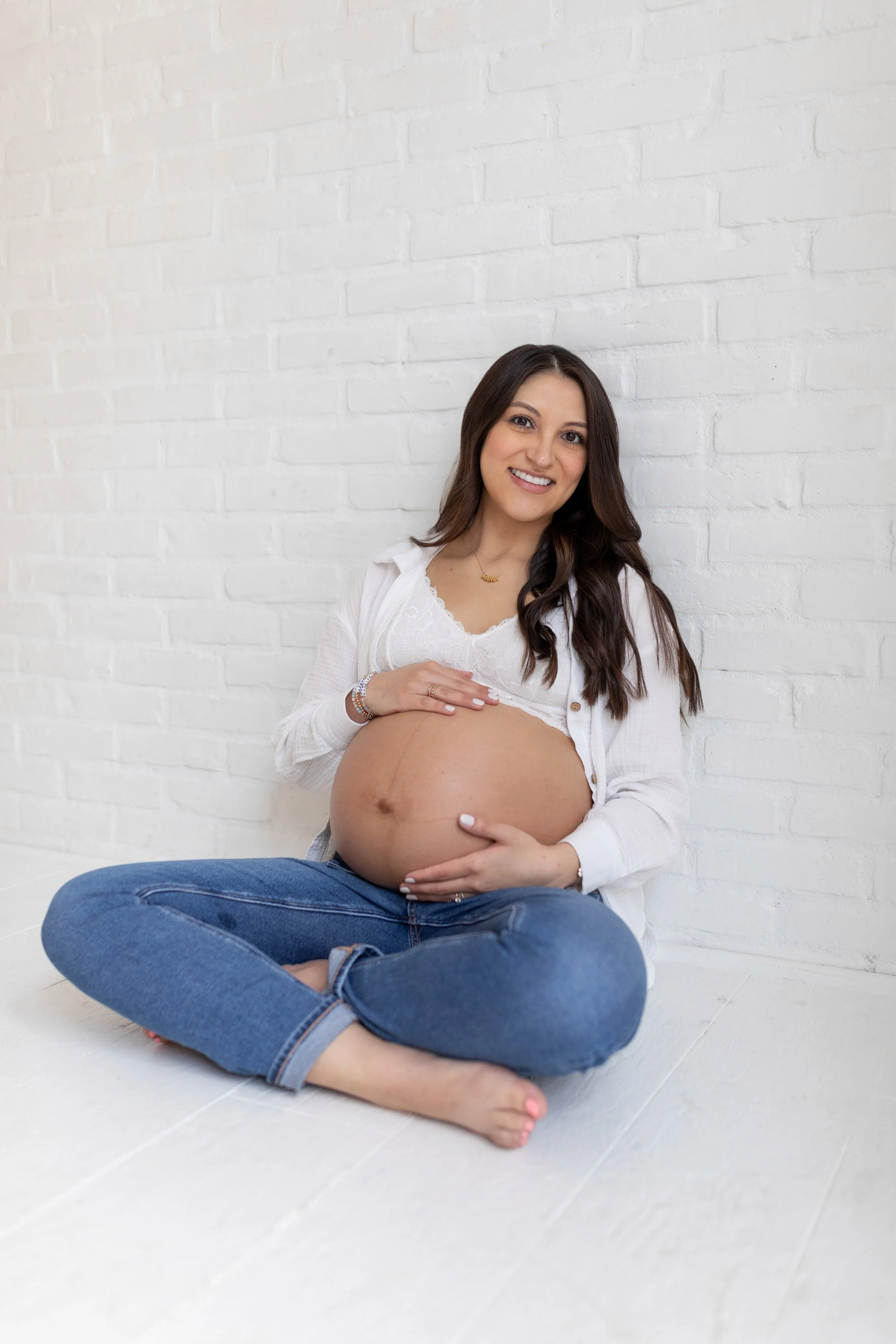 A pregnant woman sitting cross-legged on a white floor, smiling, with a white brick wall behind her.