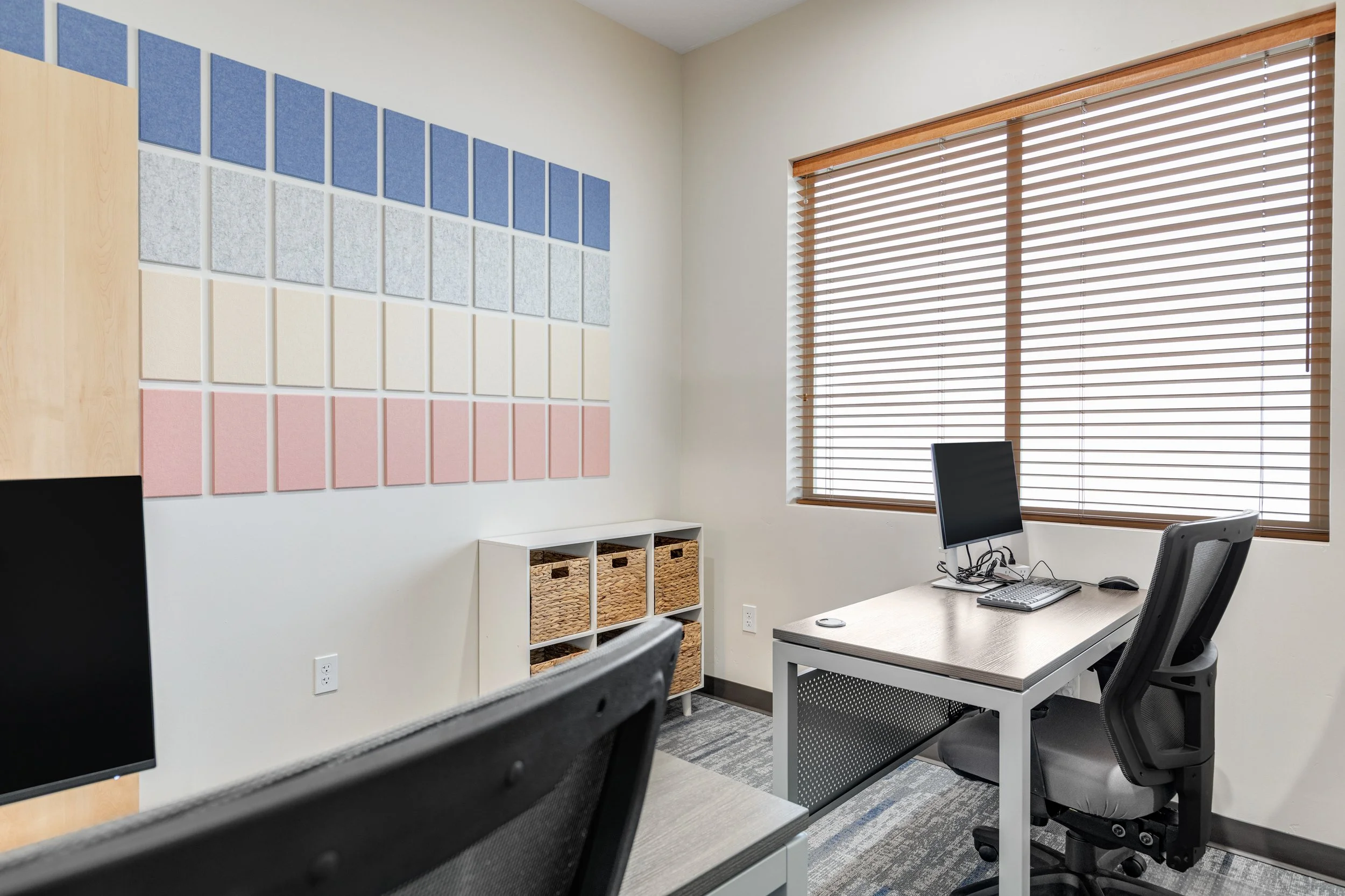 Office room with a desk, computer, and a window with blinds. Wall art with colored soundproofing panels.