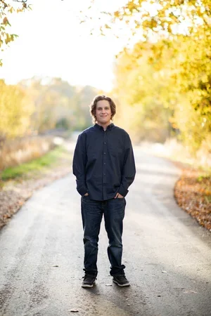 A young man standing on a rural road surrounded by autumn trees with yellow and orange leaves, wearing a dark blue shirt and jeans.