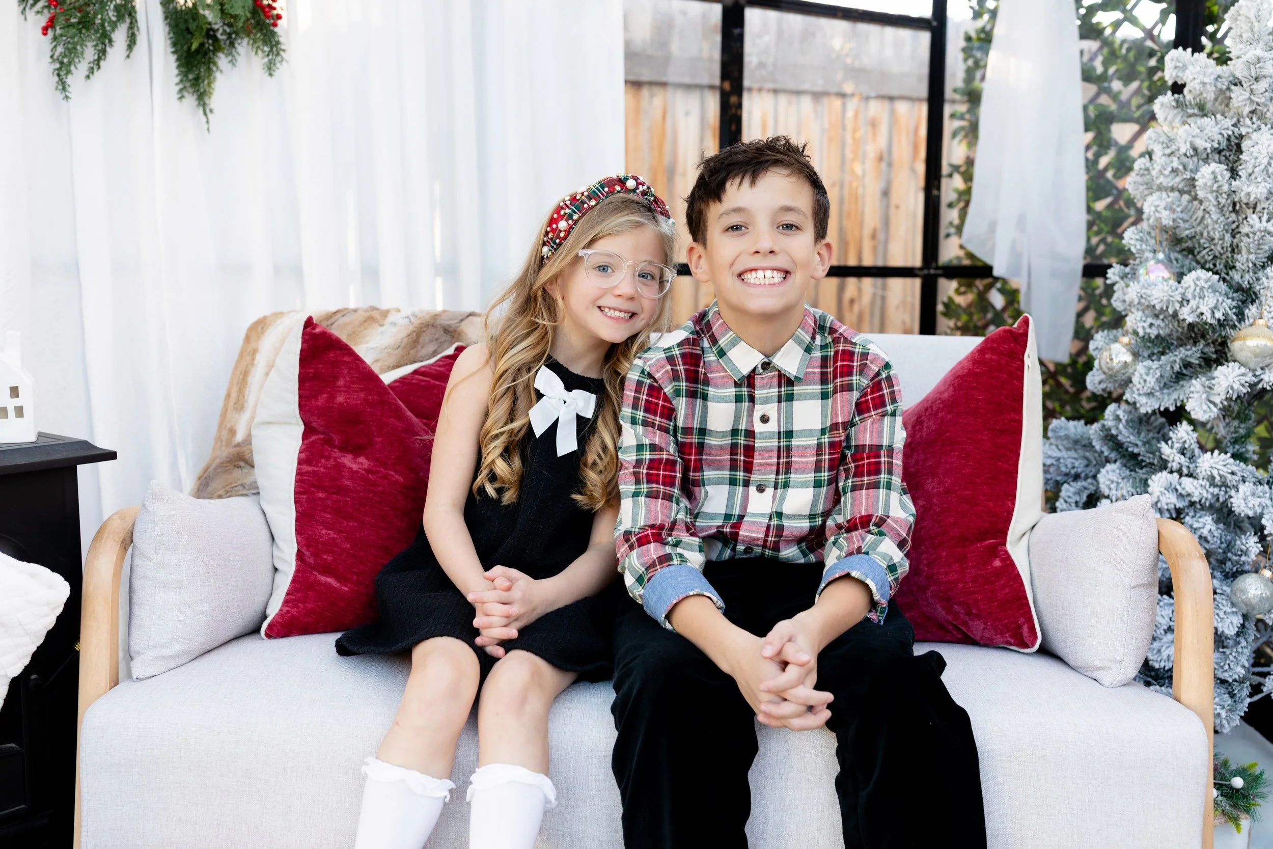 Two children sitting on a white sofa, smiling, with a Christmas tree and holiday decorations in the background.