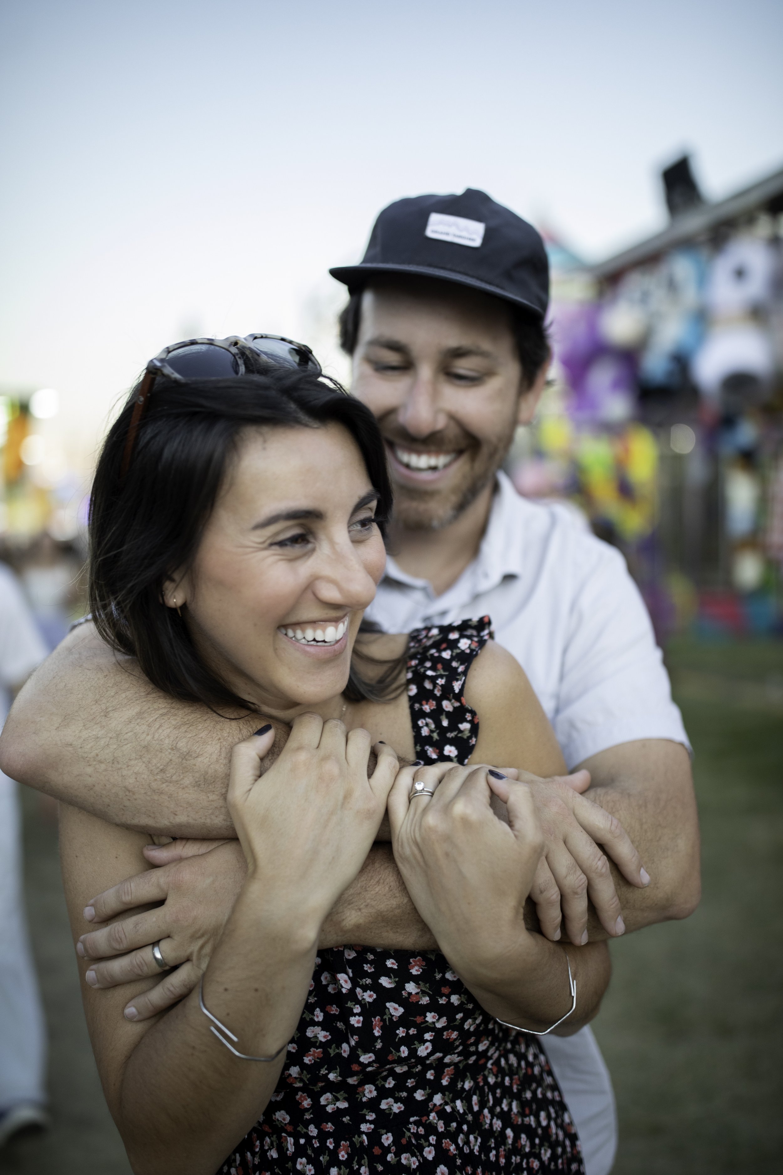 A man and woman embracing and smiling at an outdoor fair or festival.