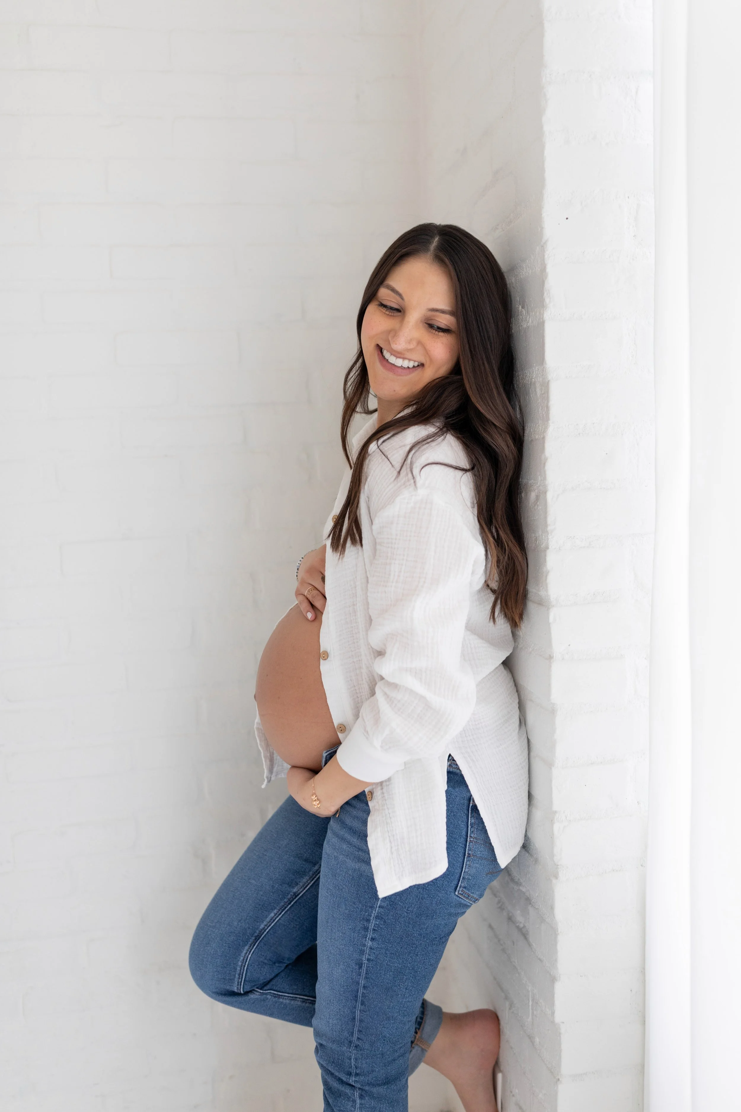 Pregnant woman smiling while leaning against a white brick wall, wearing a white shirt and jeans.