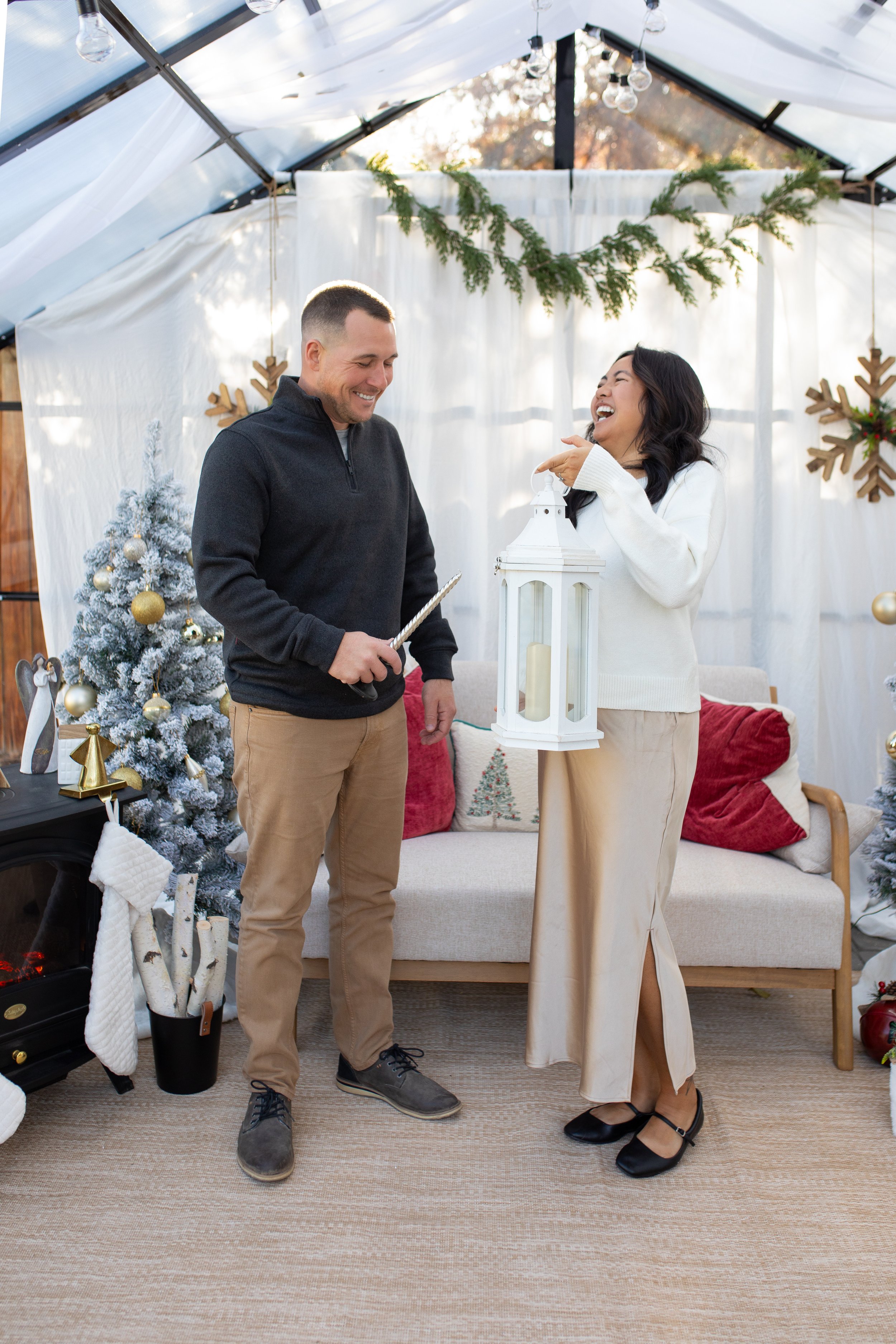 A couple is in a decorated room with holiday decorations, including Christmas trees and hanging snowflakes. They are smiling and laughing, with one person holding a lantern and the other holding a wand, suggesting a playful holiday activity.