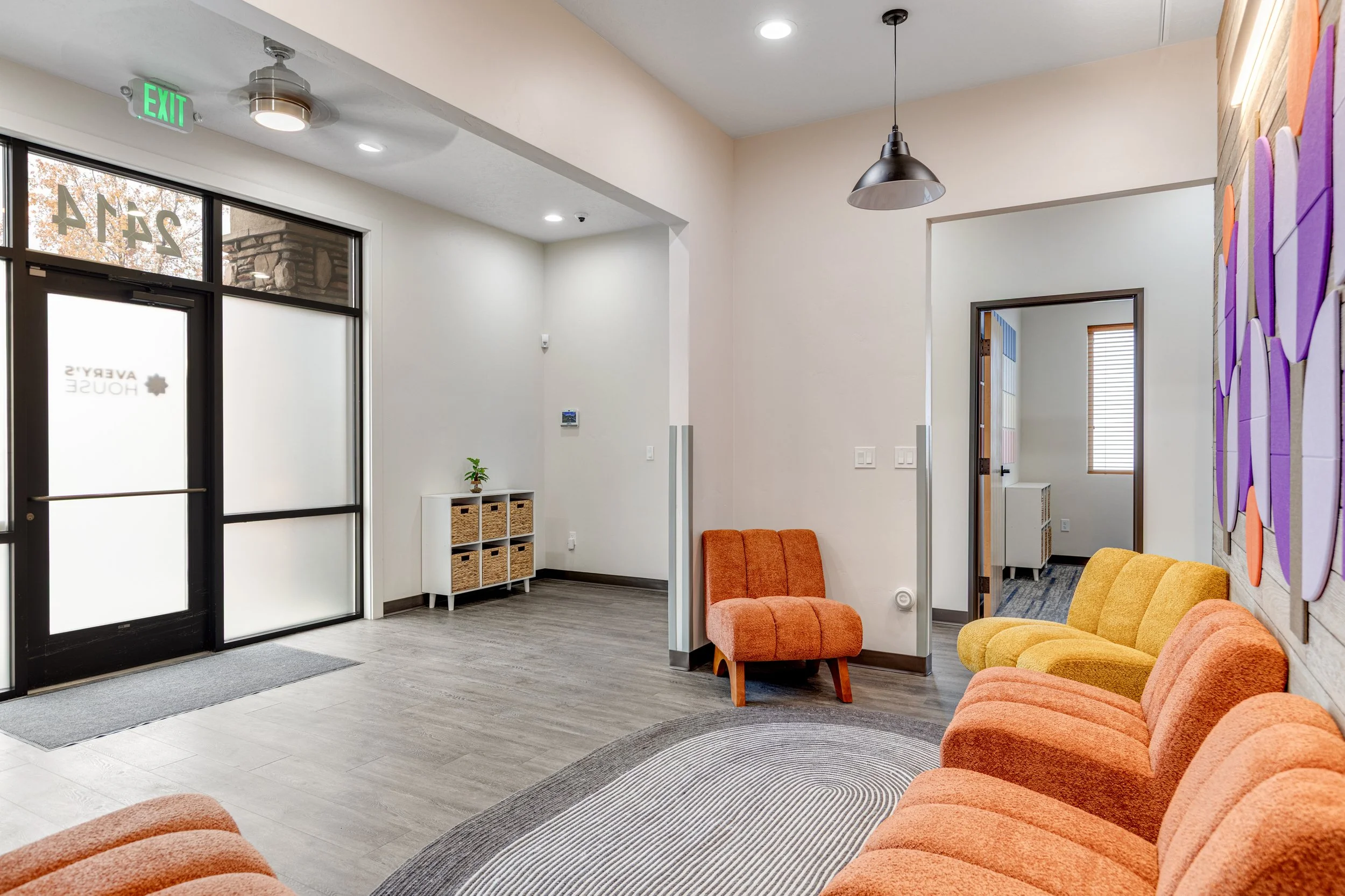 Lobby with orange and yellow upholstered chairs, a gray and white circular rug, a small shelf with wicker baskets, and a glass door with frosted and clear glass panels, leading outside. There is a doorway to another room, a ceiling fan, and a hanging