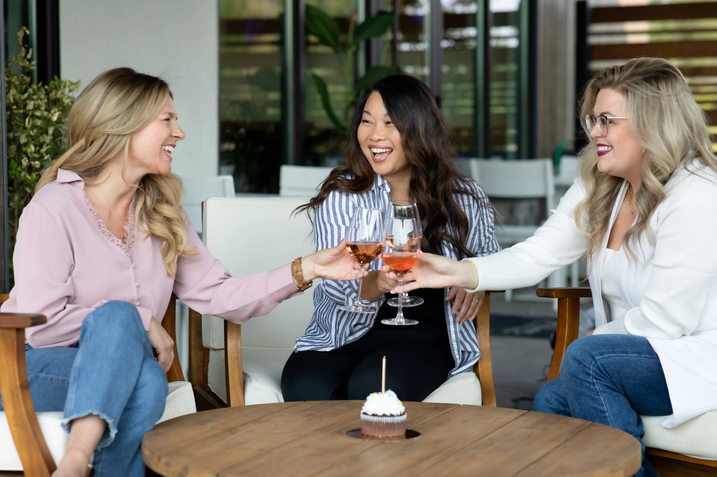Three women celebrating with glasses of rosé wine around a birthday cake on a table.