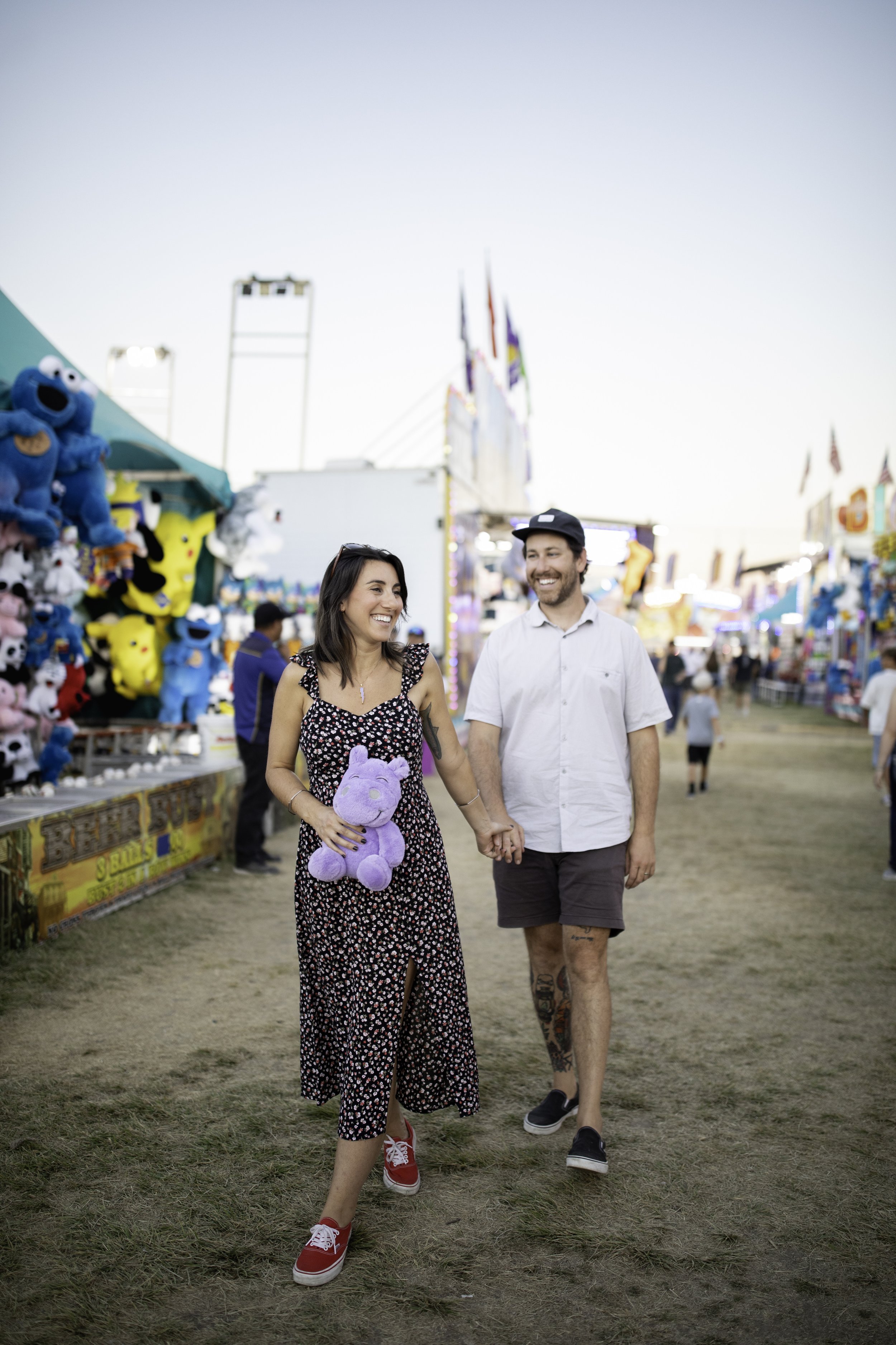 A smiling couple walking hand-in-hand at a fairground with game booths and prizes, including plush toys, in the background.