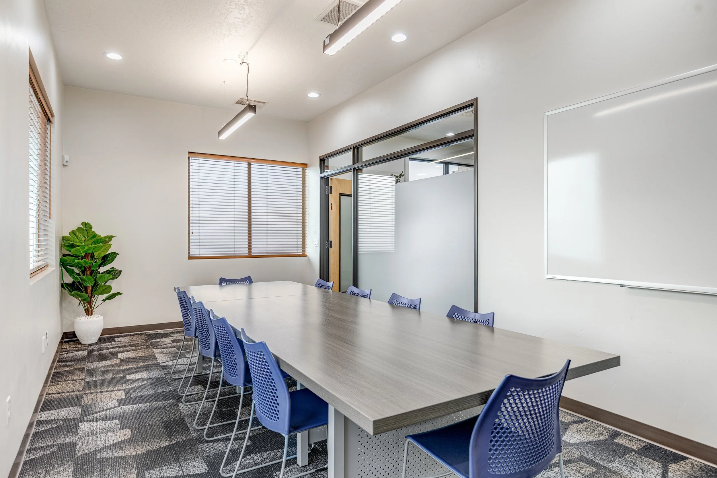 Empty conference room with a long table, blue chairs, large windows with blinds, a potted plant, a whiteboard, and glass walls.