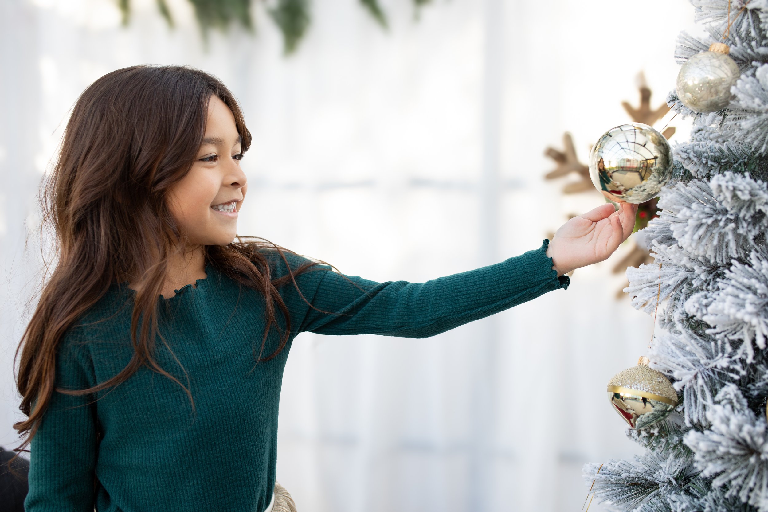A young girl with long brown hair wearing a teal sweater decorates a frosted Christmas tree with gold and silver ornaments.