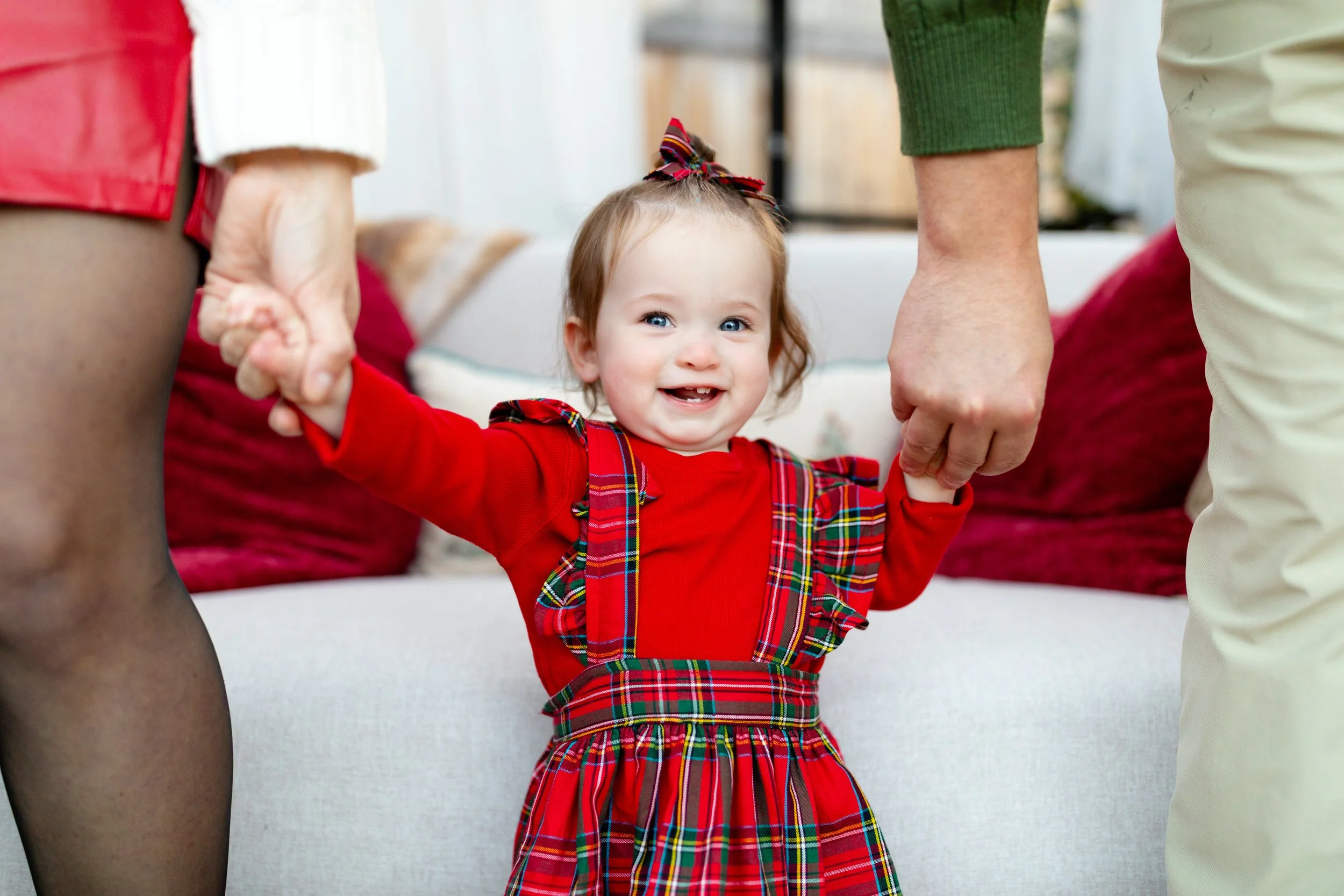 A young girl in a red plaid dress with suspenders is smiling while holding hands with two adults.