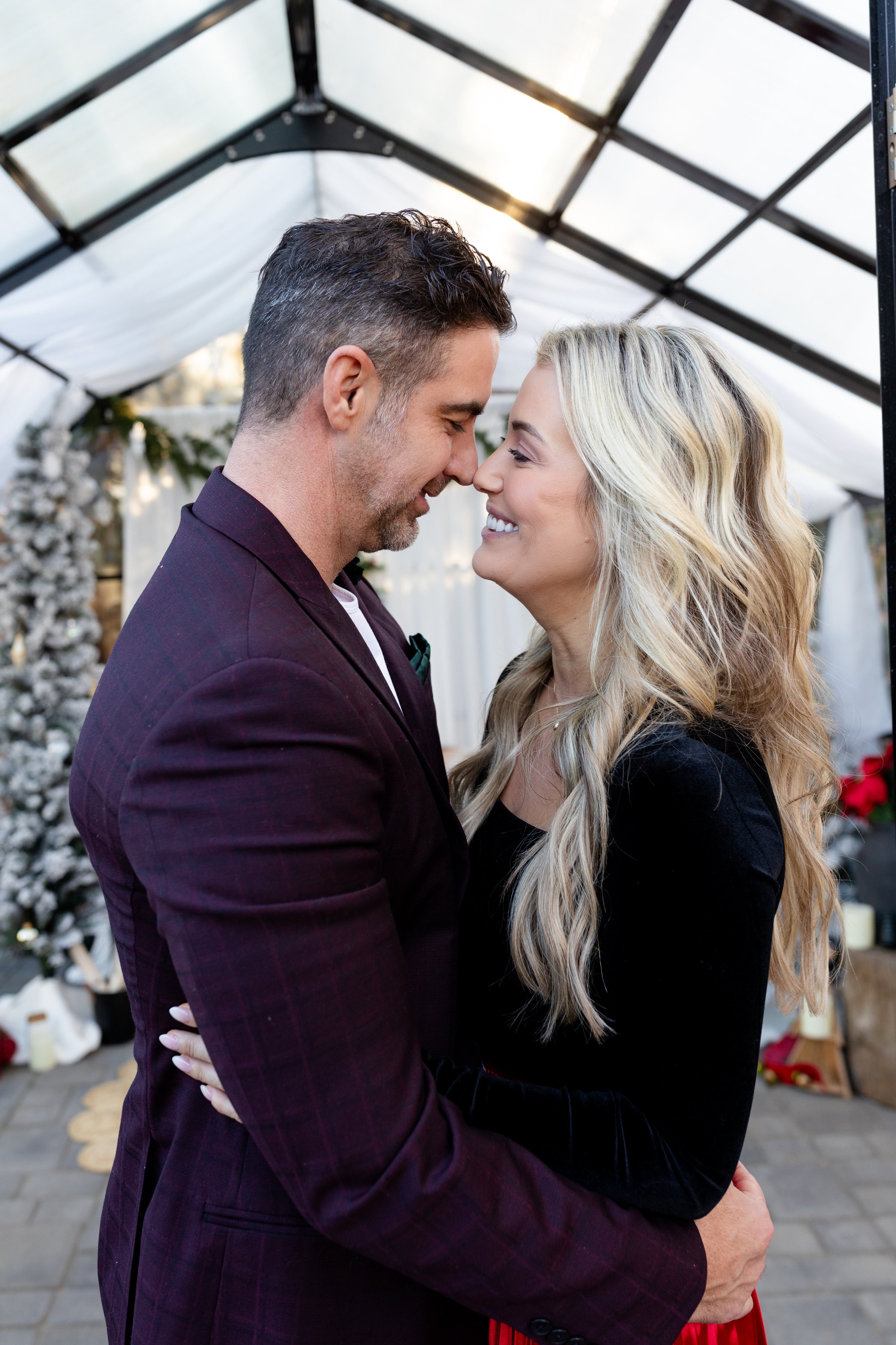 A couple is smiling and close together inside a decorated greenhouse or tent, with Christmas trees and holiday decorations in the background.
