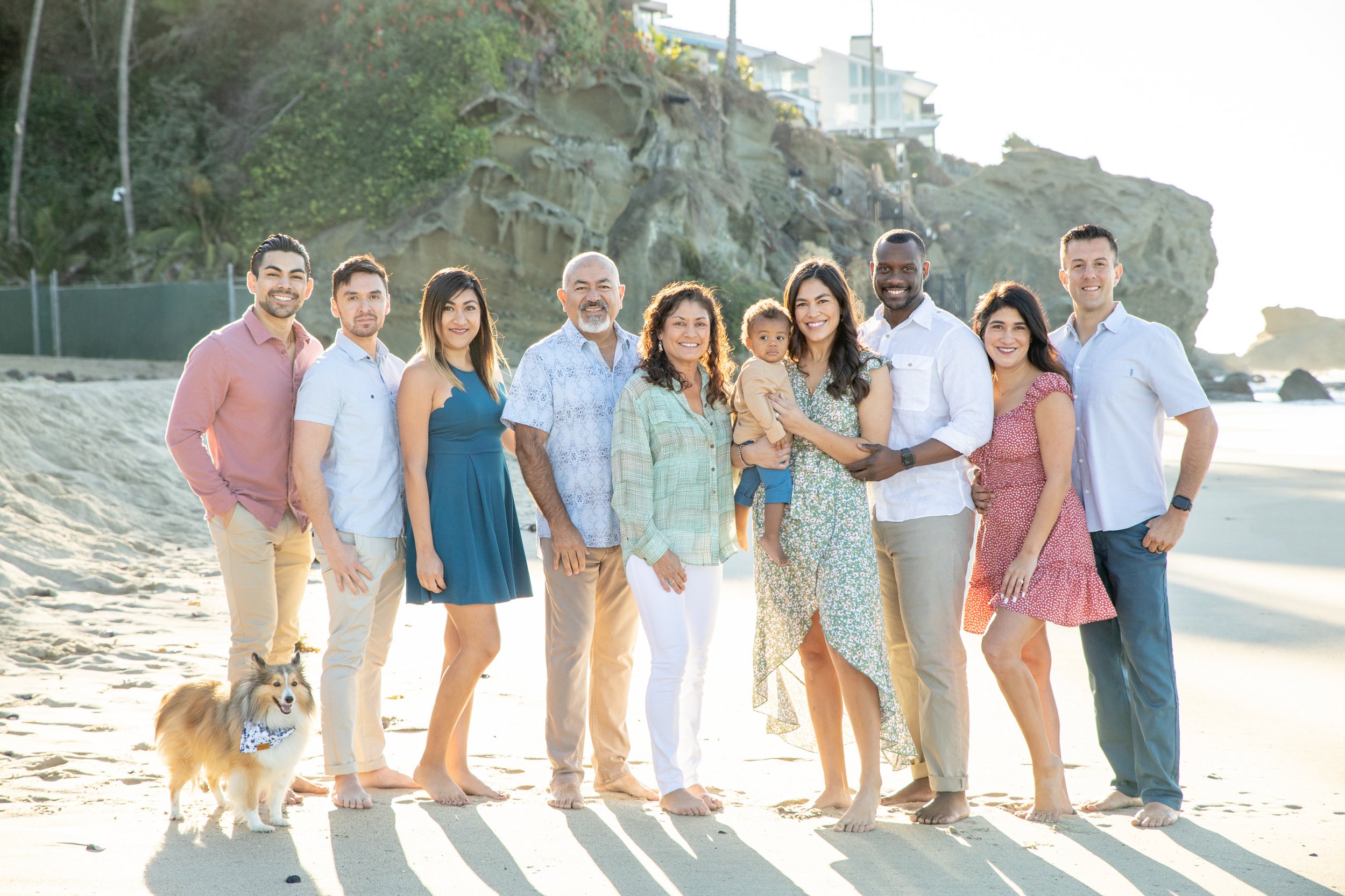A diverse group of ten people, including children and adults, along with a dog, pose on a beach with rocky cliffs and ocean in the background during sunset.
