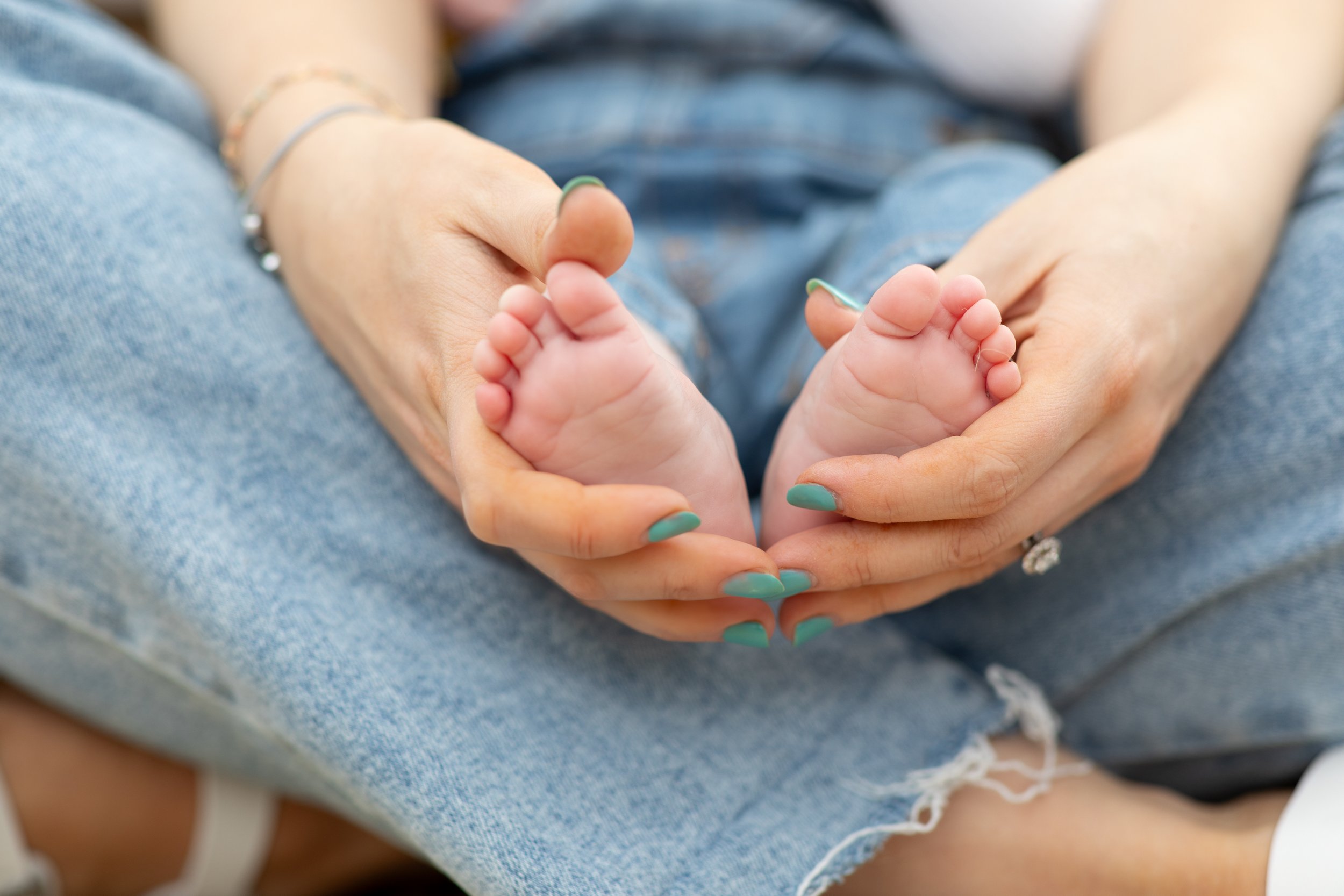 Close-up of an adult woman holding newborn baby's feet in her hands, sitting in blue jeans.