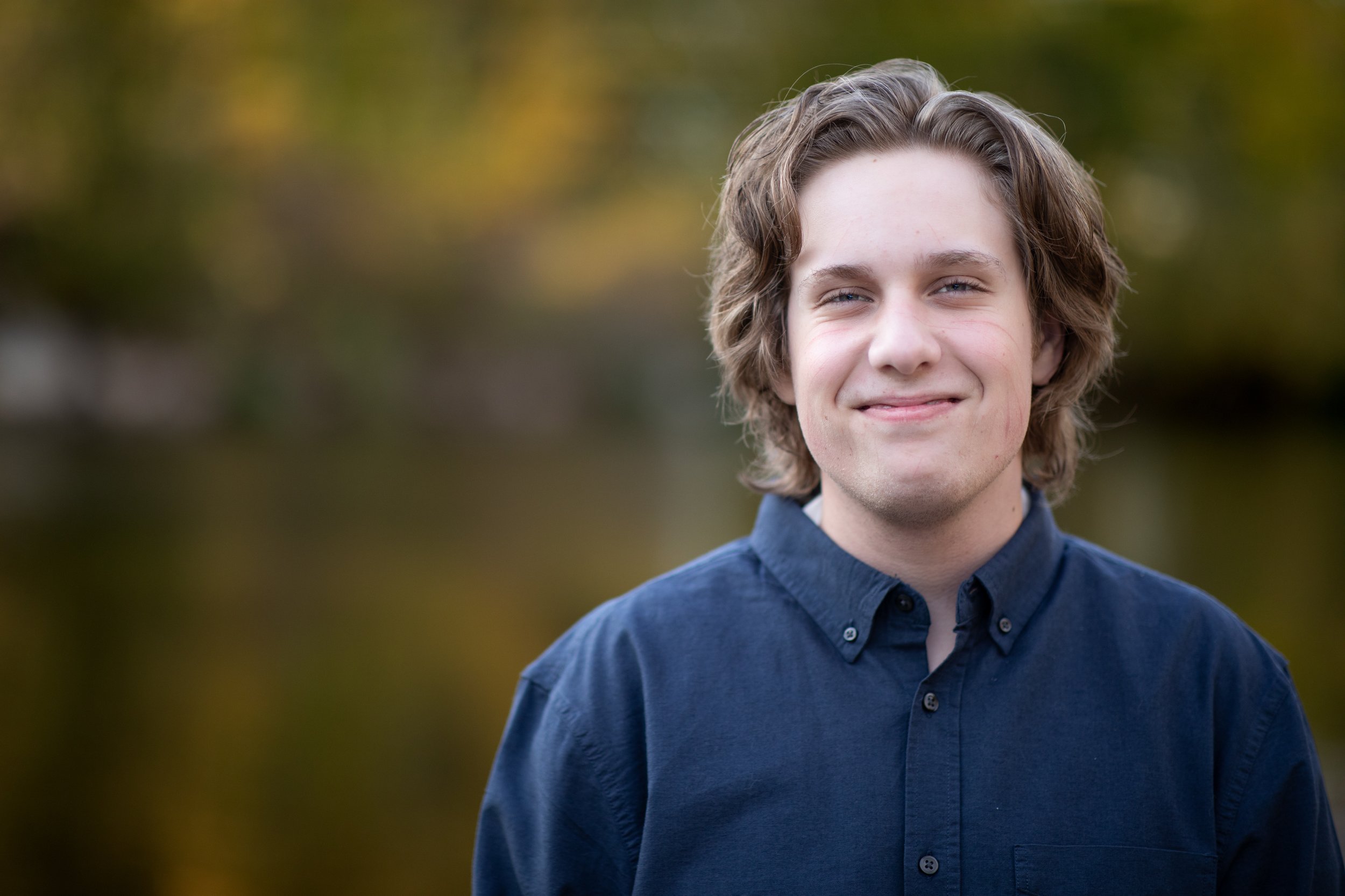 A young man with wavy brown hair, wearing a dark blue button-up shirt, standing outdoors with a blurred background of fall foliage.