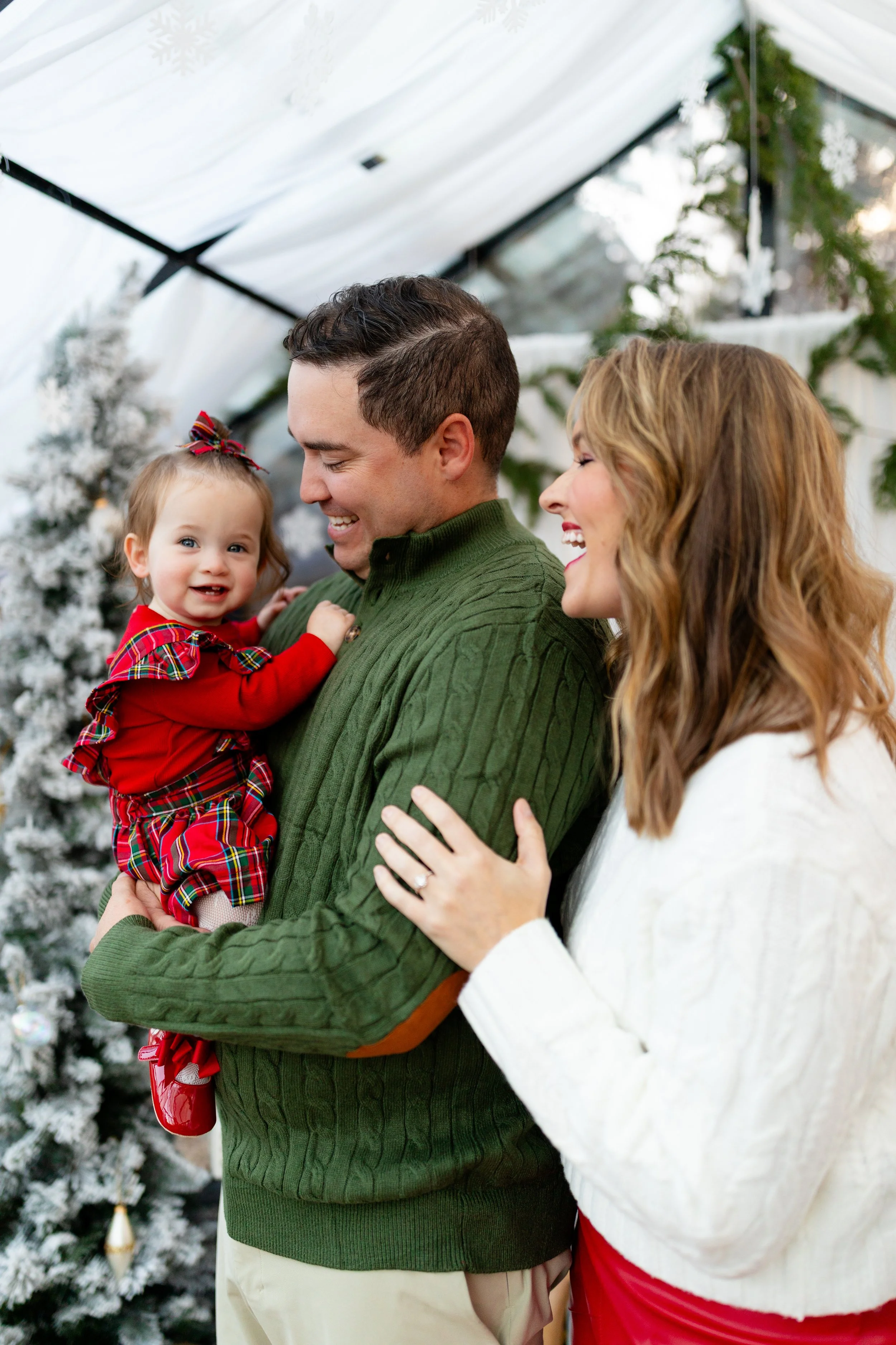 A smiling young family celebrating Christmas indoors with a decorated snowy Christmas tree in the background. The man holds a little girl dressed in red with a plaid skirt, while a woman with wavy hair stands beside them, touching the man's arm.