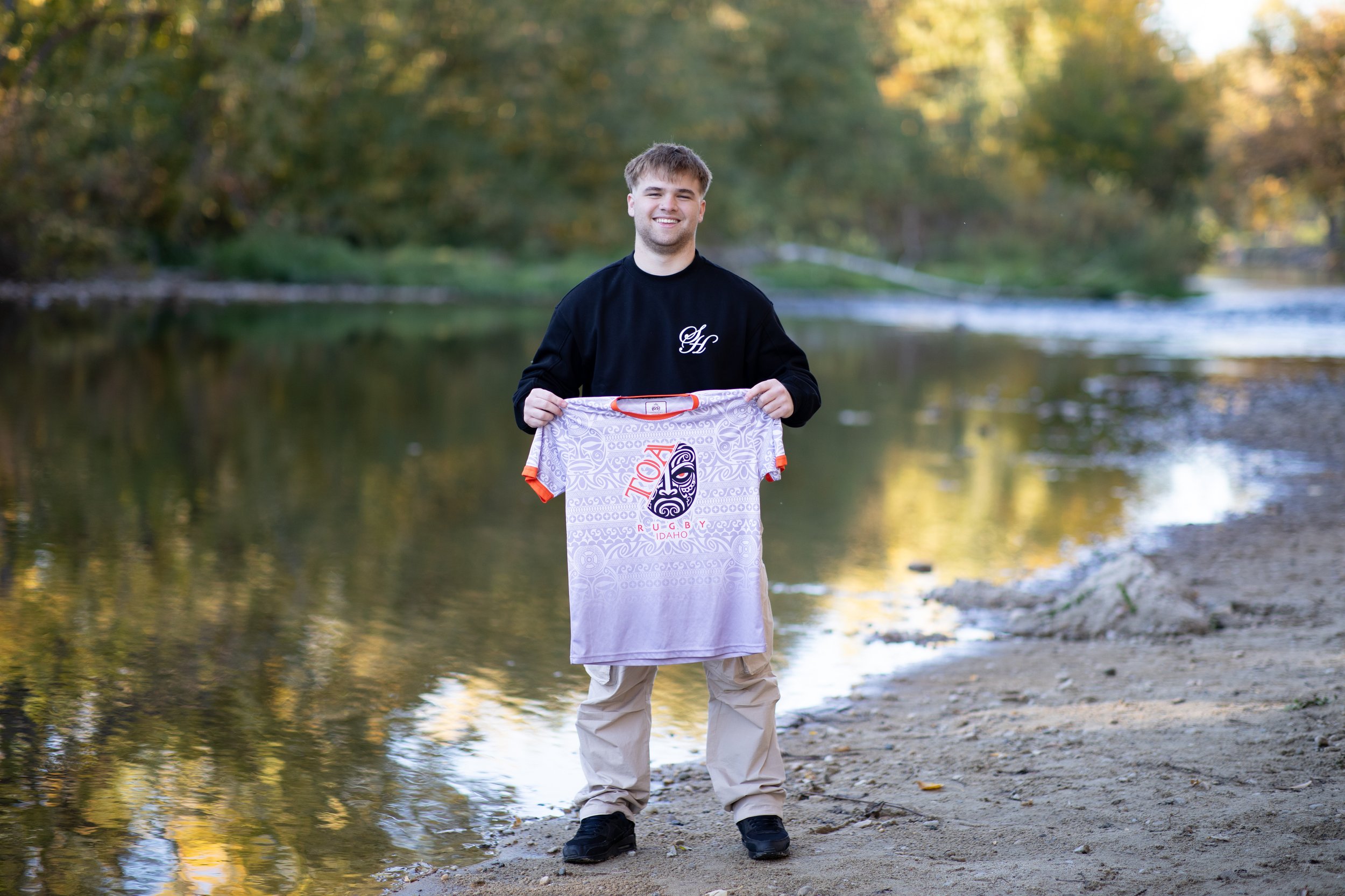 A young man standing on a riverbank holding an Idaho rugby T-shirt with a tribal mask design, wearing a black sweatshirt and beige pants, smiling with trees and water in the background.