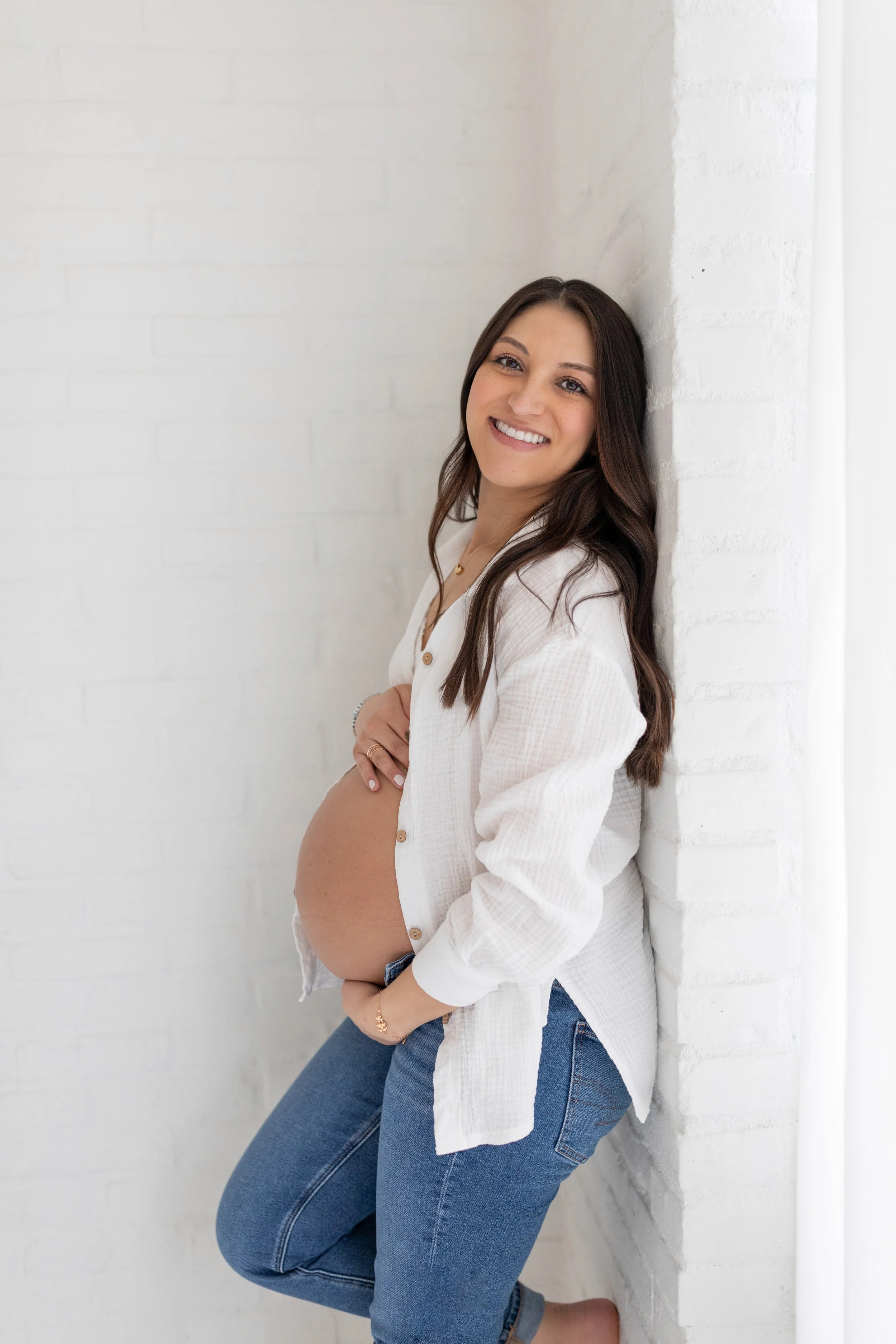 A pregnant woman with long brown hair smiling while leaning against a white brick wall in a white button-up shirt and blue jeans.