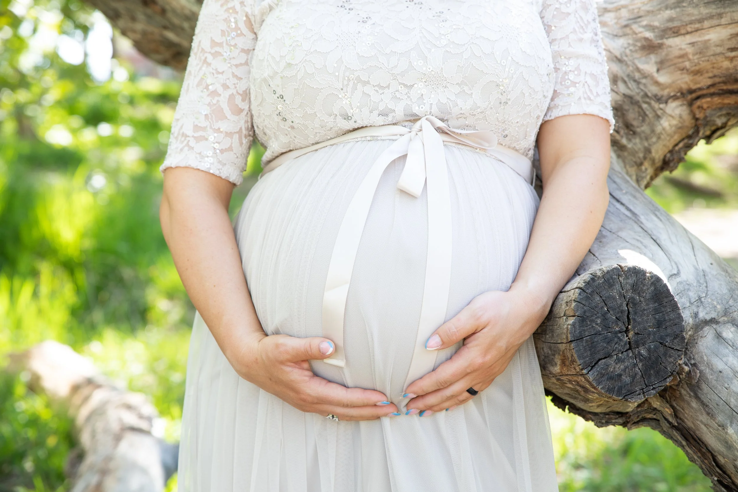 Close-up of a pregnant woman in a white lace dress, cradling her belly with both hands outdoors against a fallen tree trunk with green foliage in the background.