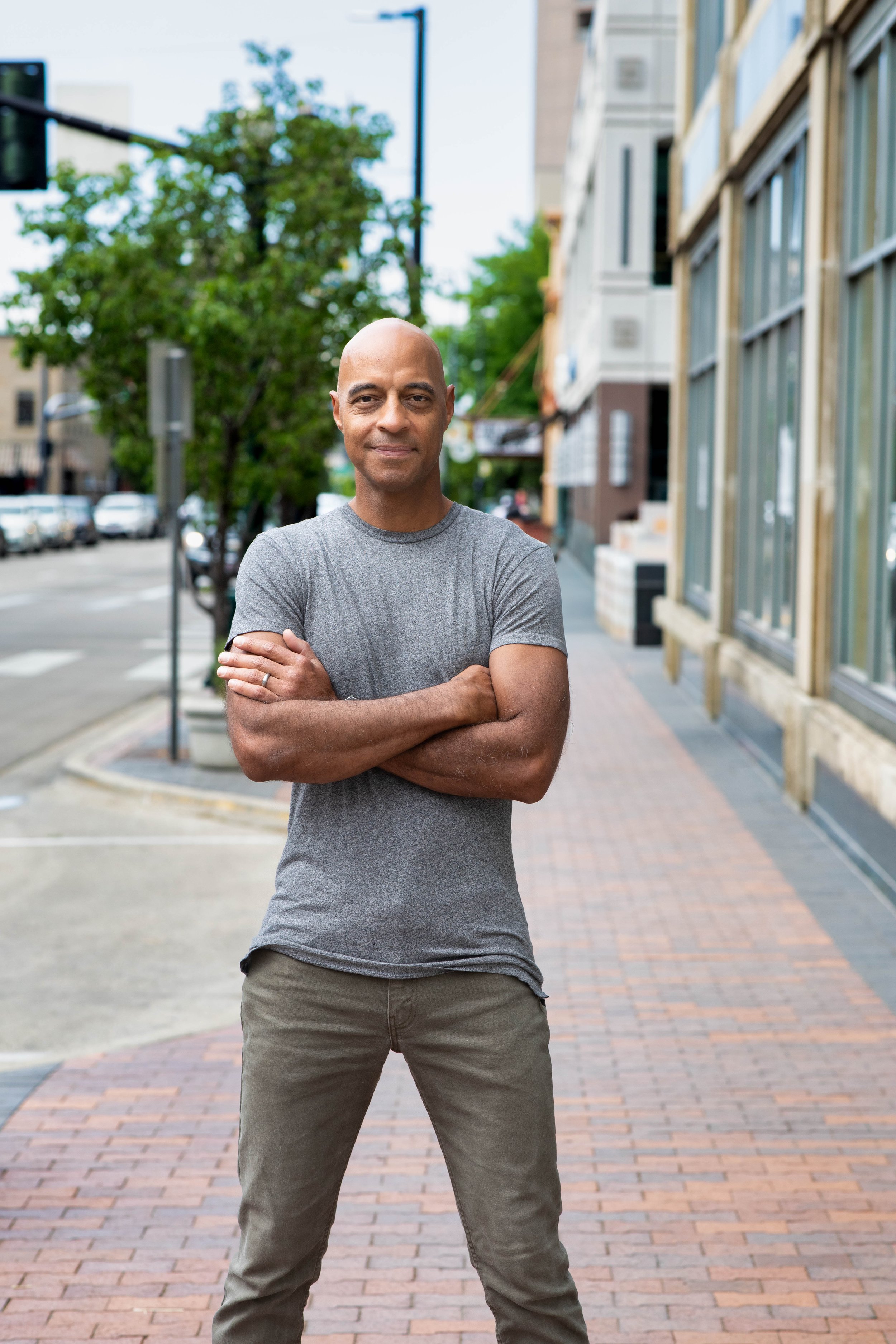A man standing with arms crossed on a brick sidewalk in an urban area with buildings and trees in the background.