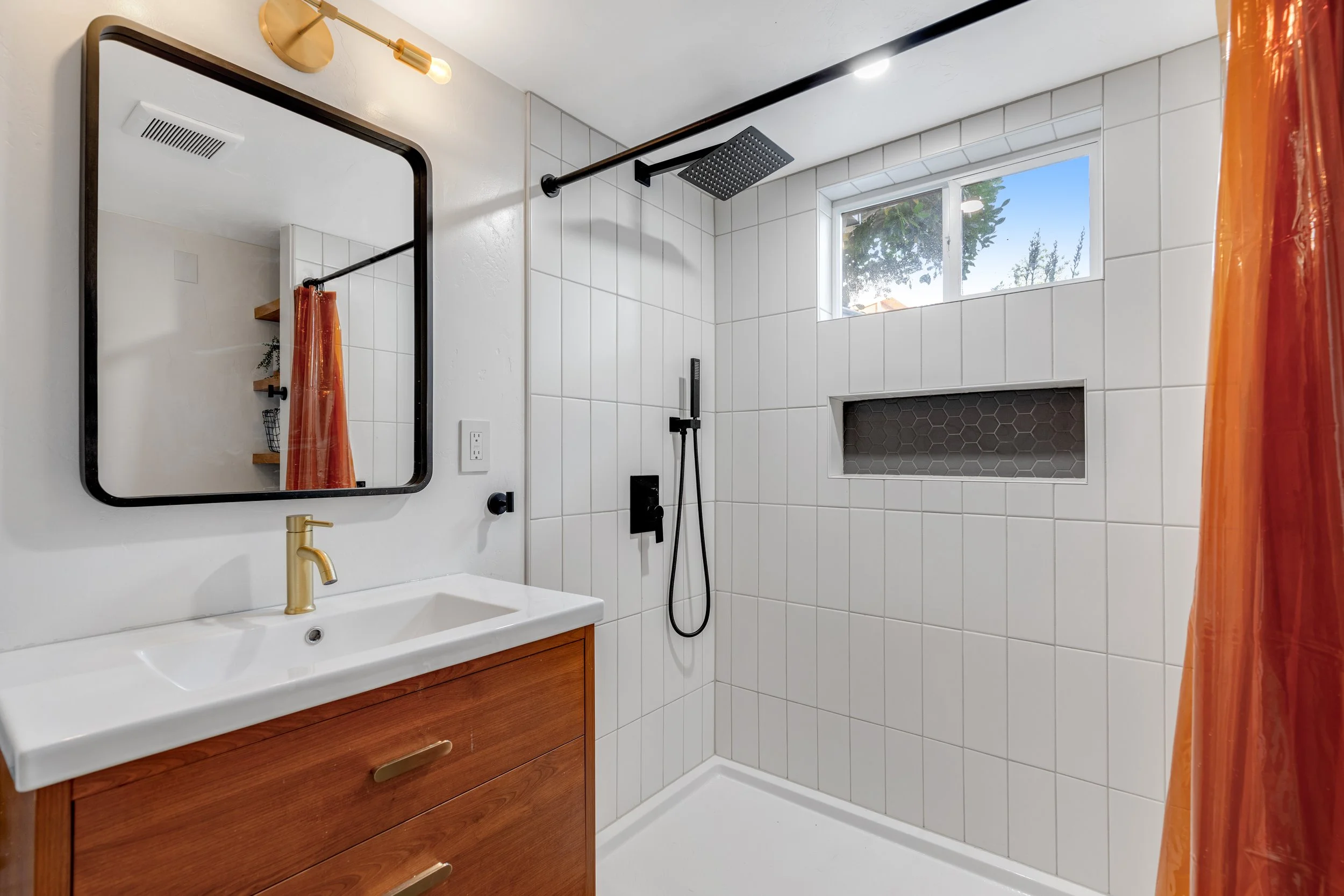 Modern bathroom with white tiled shower area, black fixtures, small window showing trees outside, wooden vanity with gold faucet, rectangular mirror with black frame, orange shower curtain, and built-in shelf with gray tiles.