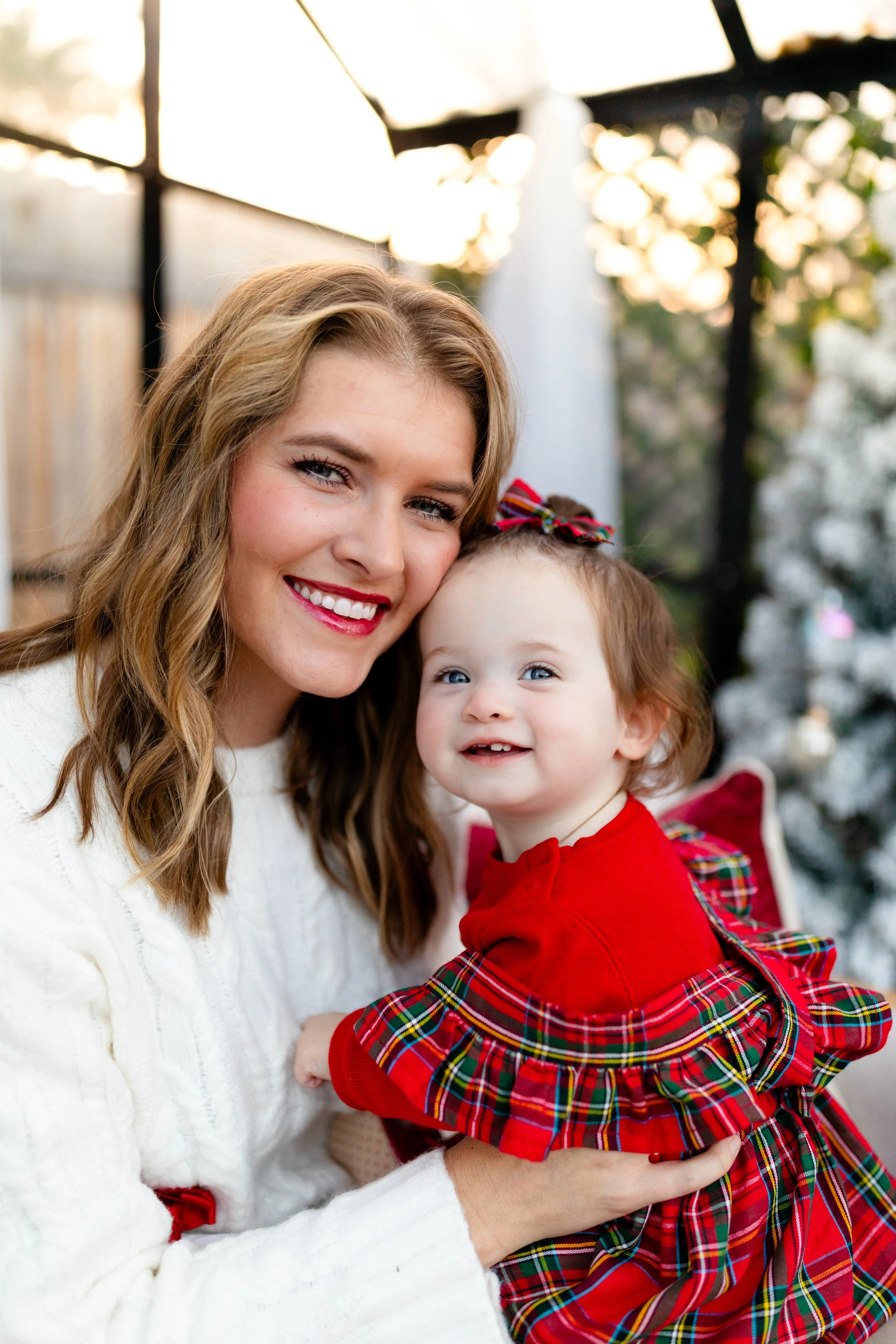 A smiling woman with light brown hair and a young girl with curly hair, wearing a red dress with plaid ruffle, sitting together outdoors near a Christmas tree.