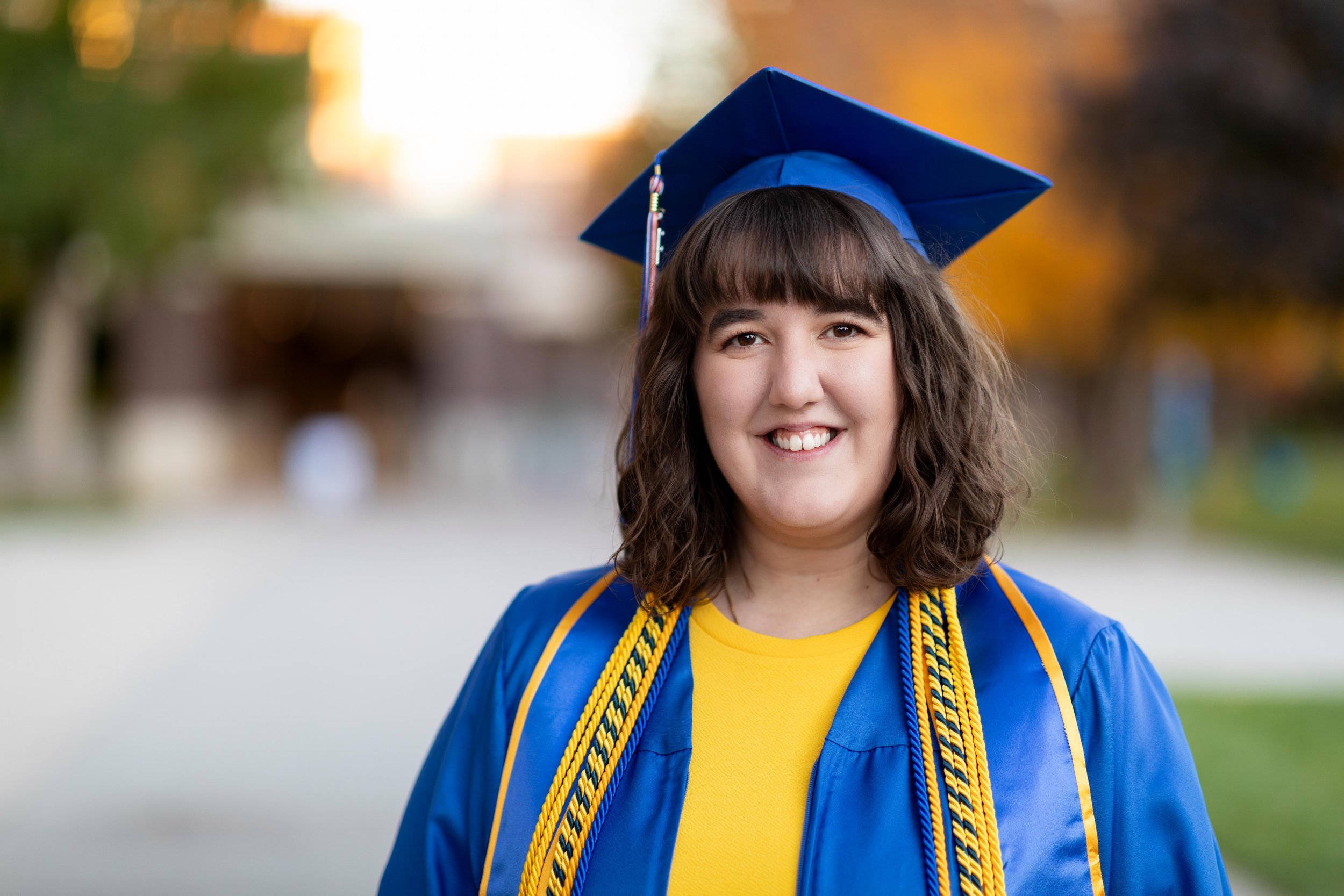 A young woman in a blue graduation cap and gown, smiling outdoors with autumn trees in the background.
