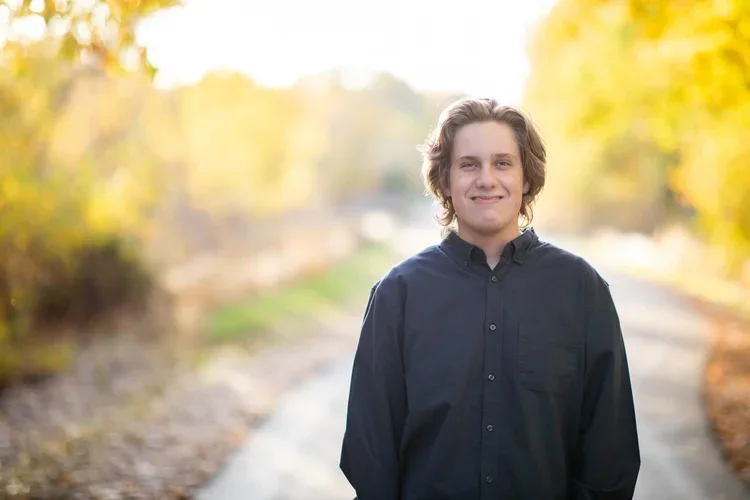 Young man with wavy hair wearing a dark button-up shirt standing outdoors on a sunny autumn day with yellow and orange trees in the background.