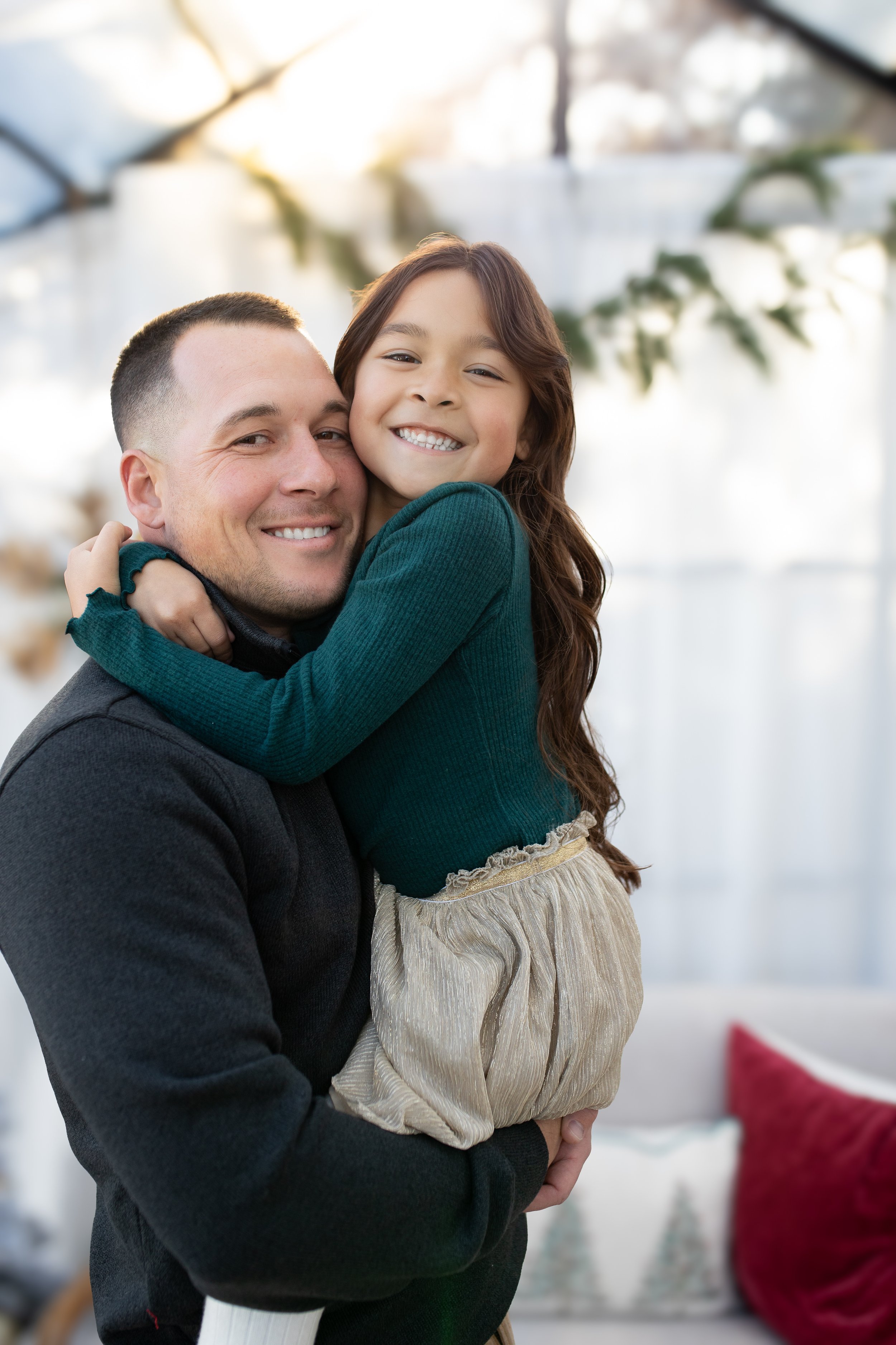 A man holding a young girl in a hug, both smiling happily indoors with a bright background and Christmas decorations.