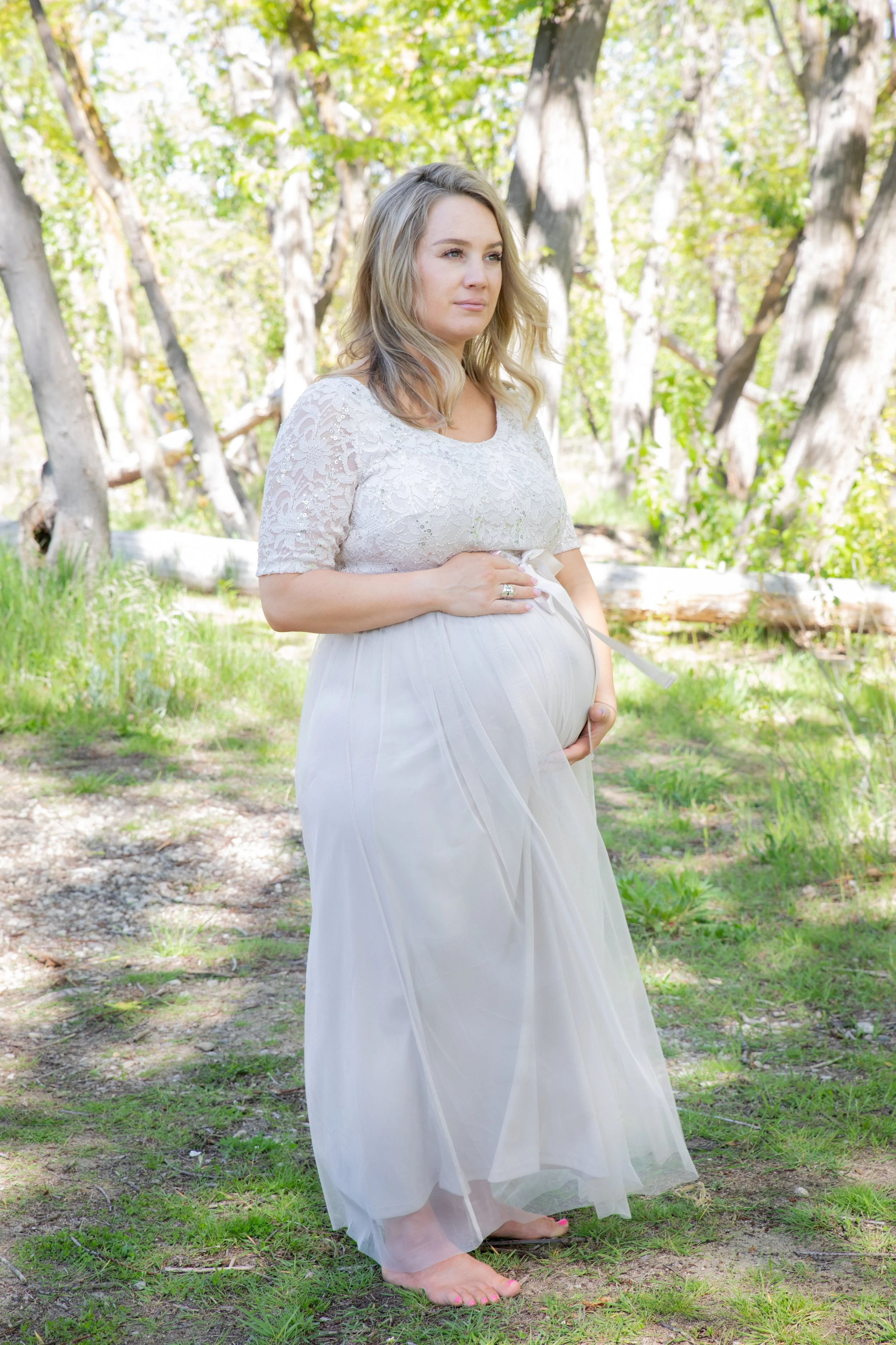 Pregnant woman standing outdoors in a forest with trees and green foliage, wearing a white lace top and a flowing white skirt, barefoot, with one hand on her belly.
