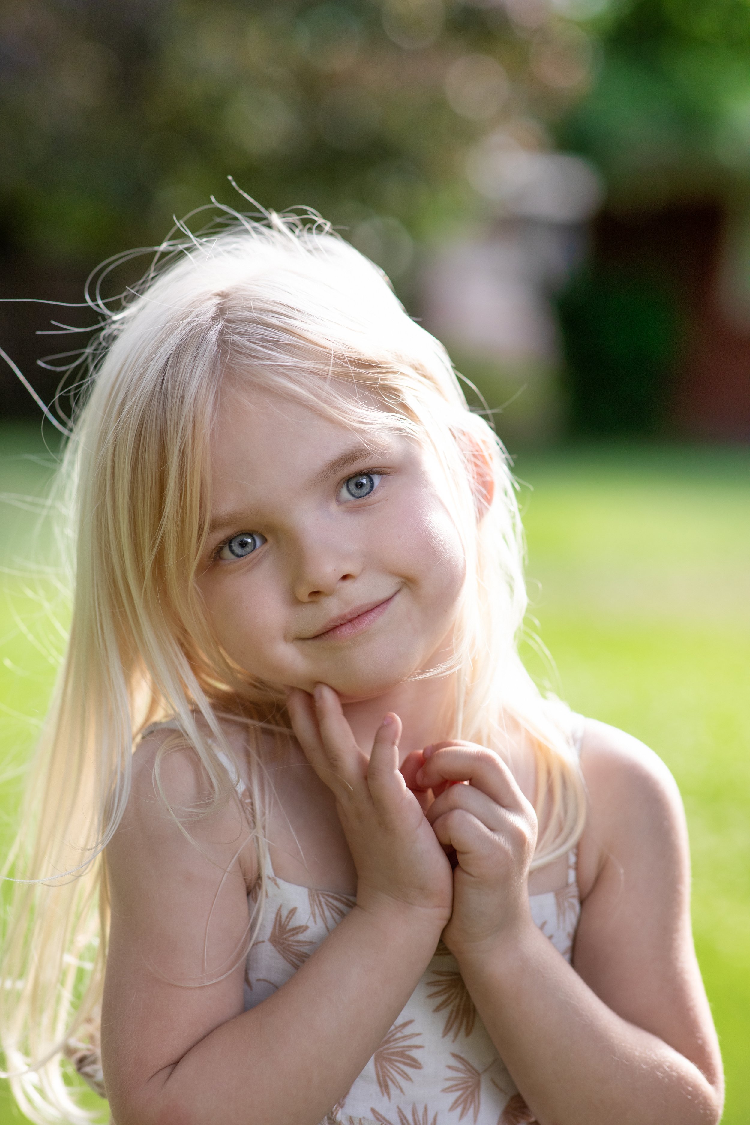 A young girl with blonde hair and blue eyes smiling outdoors in sunlight, holding her chin with one hand.