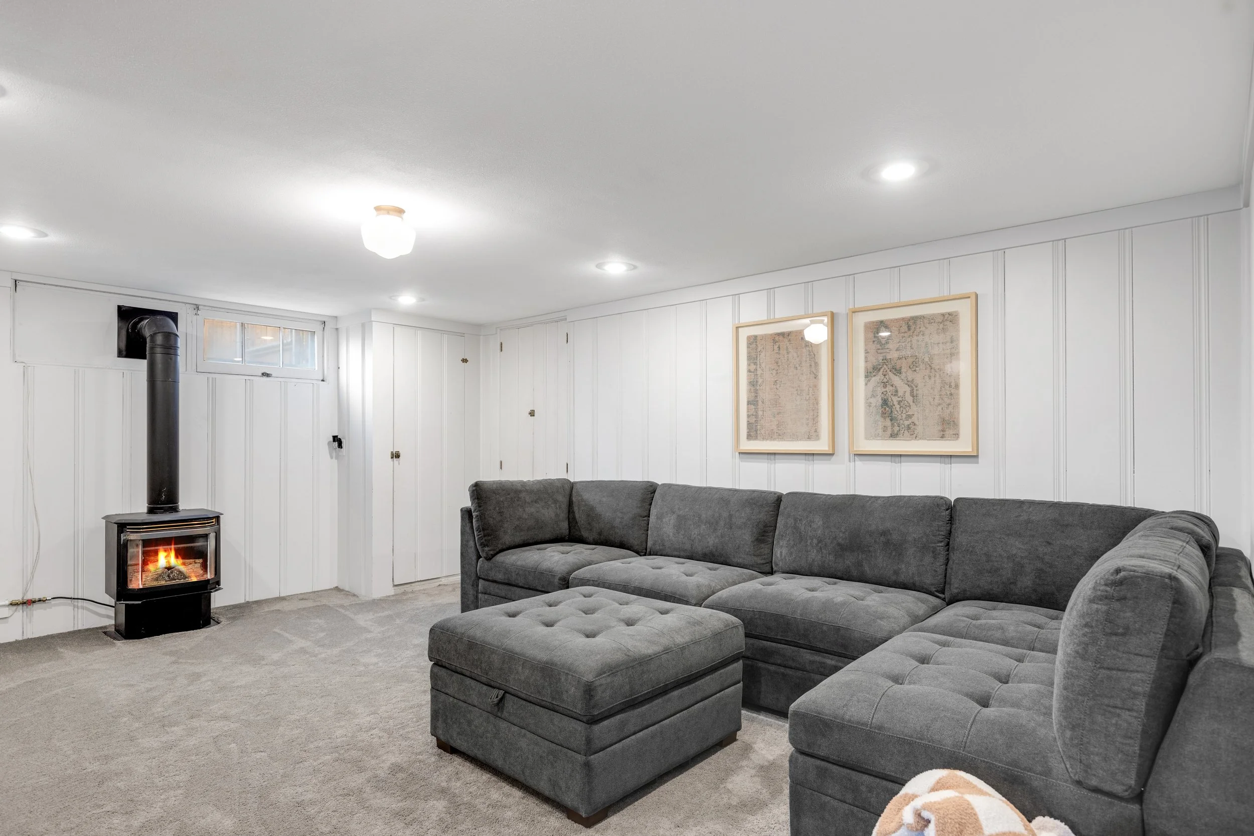 A living room with white paneled walls, a gray sectional sofa with an ottoman, a small window, a black wood-burning stove with a fire inside, and framed artwork on the wall.