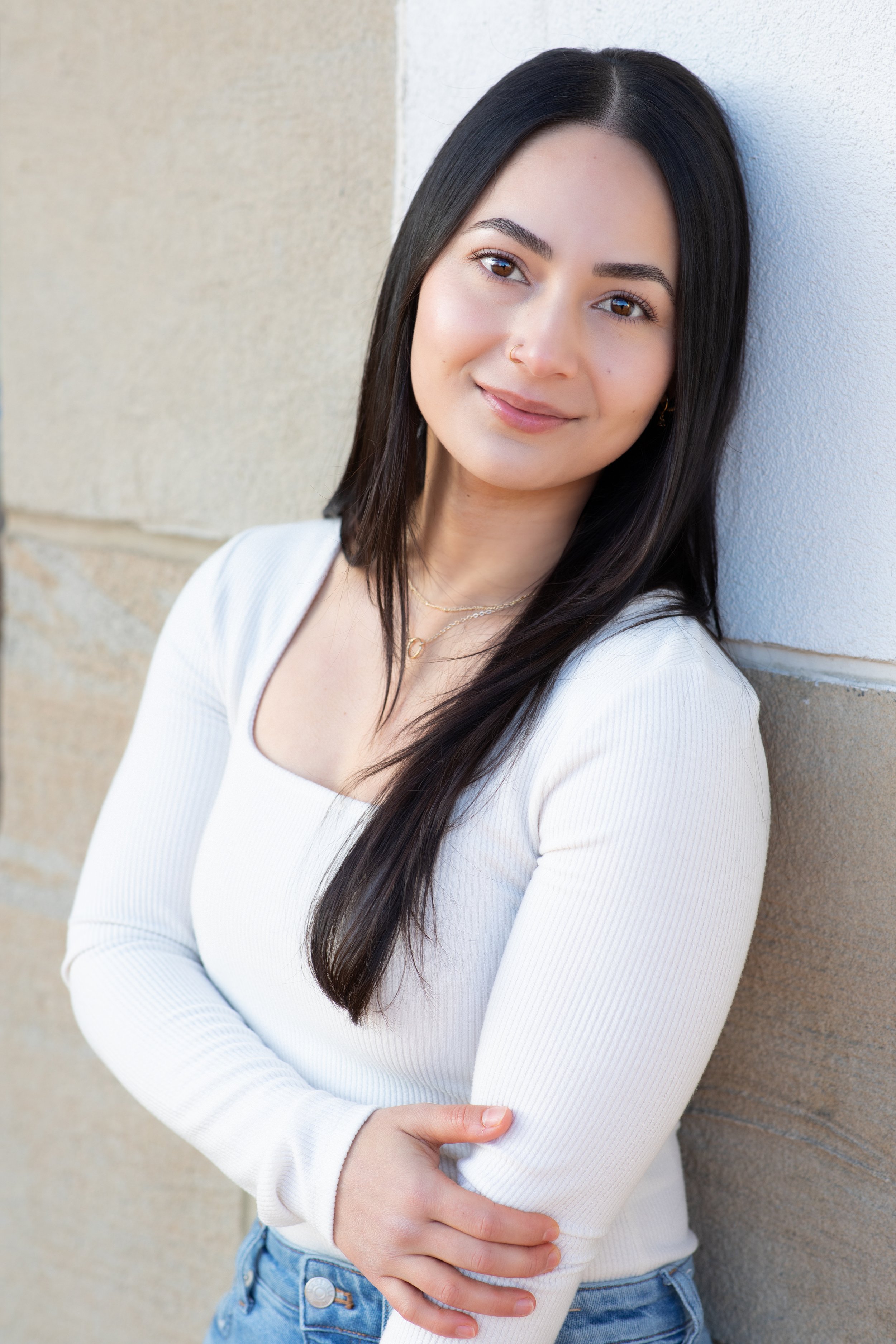 A young woman with long dark hair, wearing a white long sleeve shirt and blue jeans, smiling and leaning against a brick wall.