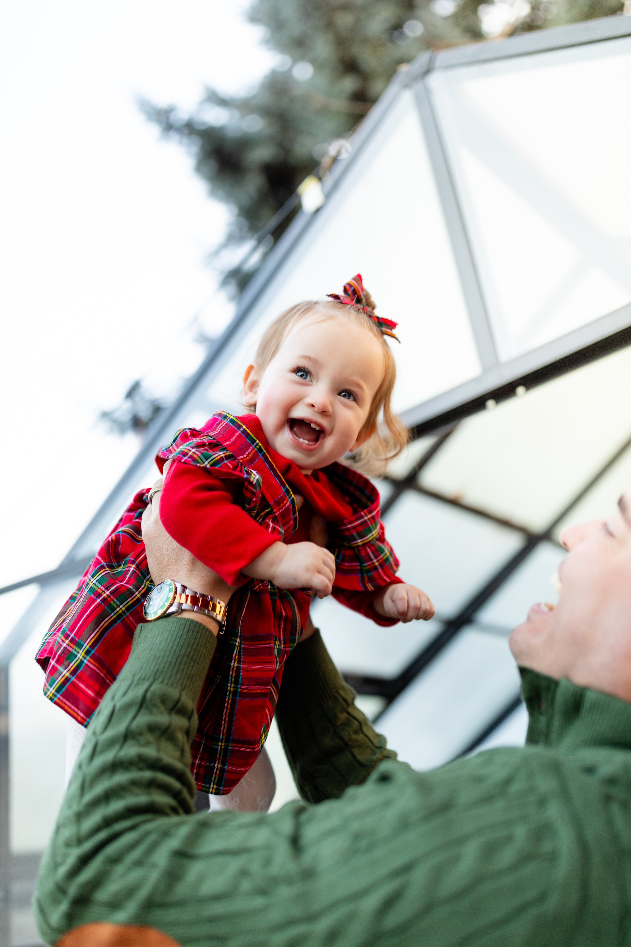 A young girl with a bow in her hair being held up by an adult, smiling and looking happy, outdoors near a solar panel.