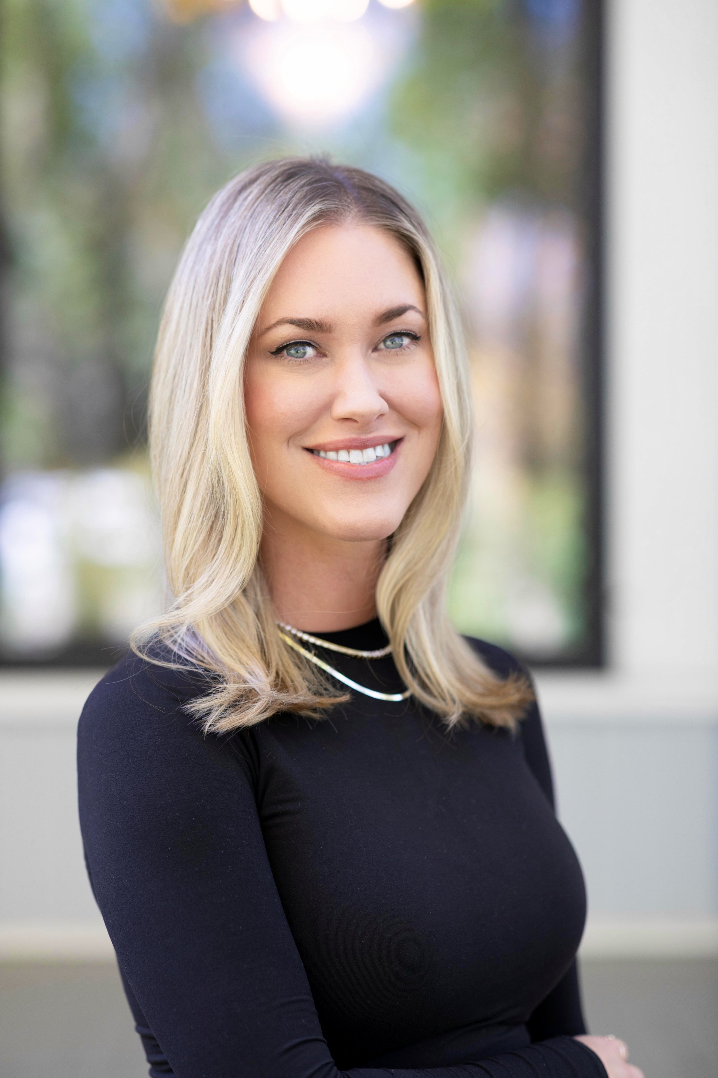 Portrait of a young woman with blonde hair and blue eyes, smiling, wearing a black top and layered necklaces, standing near a window with a blurred outdoor background.