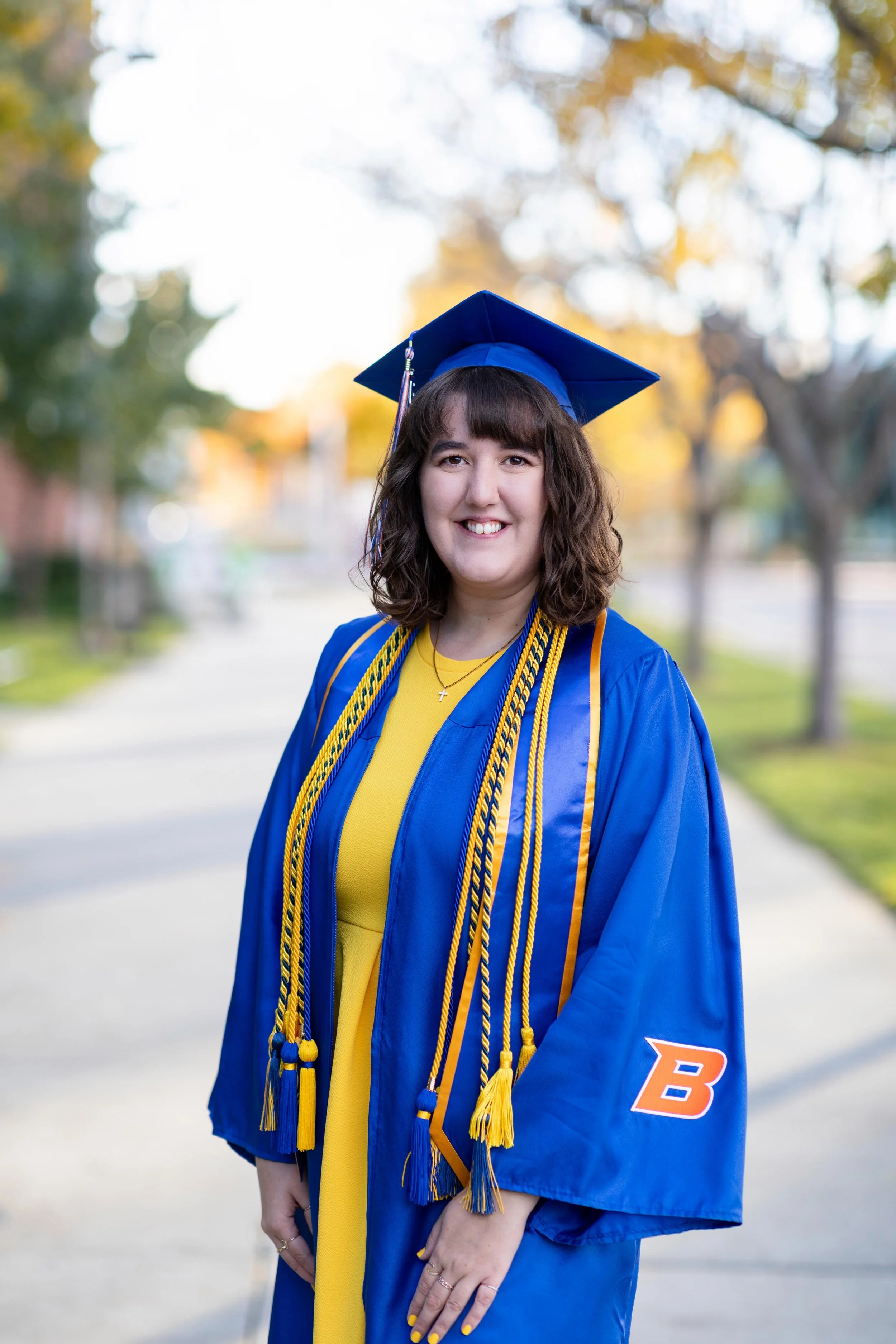A young woman in a blue graduation gown and cap, standing outdoors on a sunny day with trees in the background, smiling at the camera.
