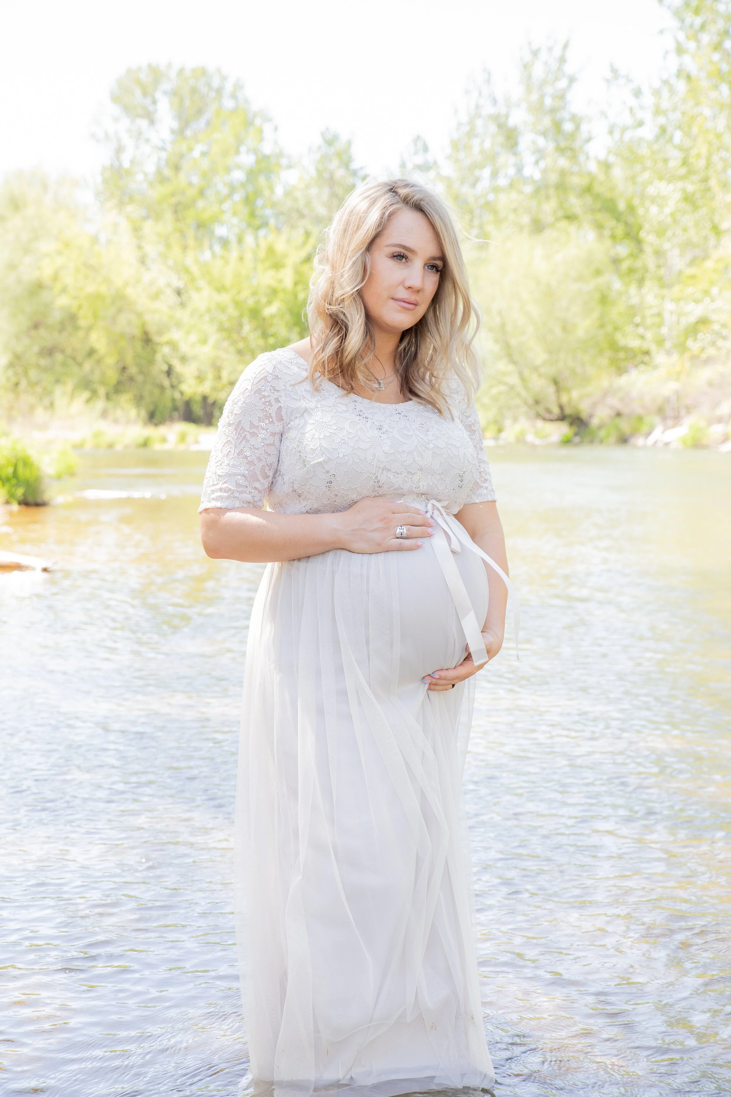 A pregnant woman in a white dress stands in a river with green trees in the background, cradling her belly and looking thoughtfully.