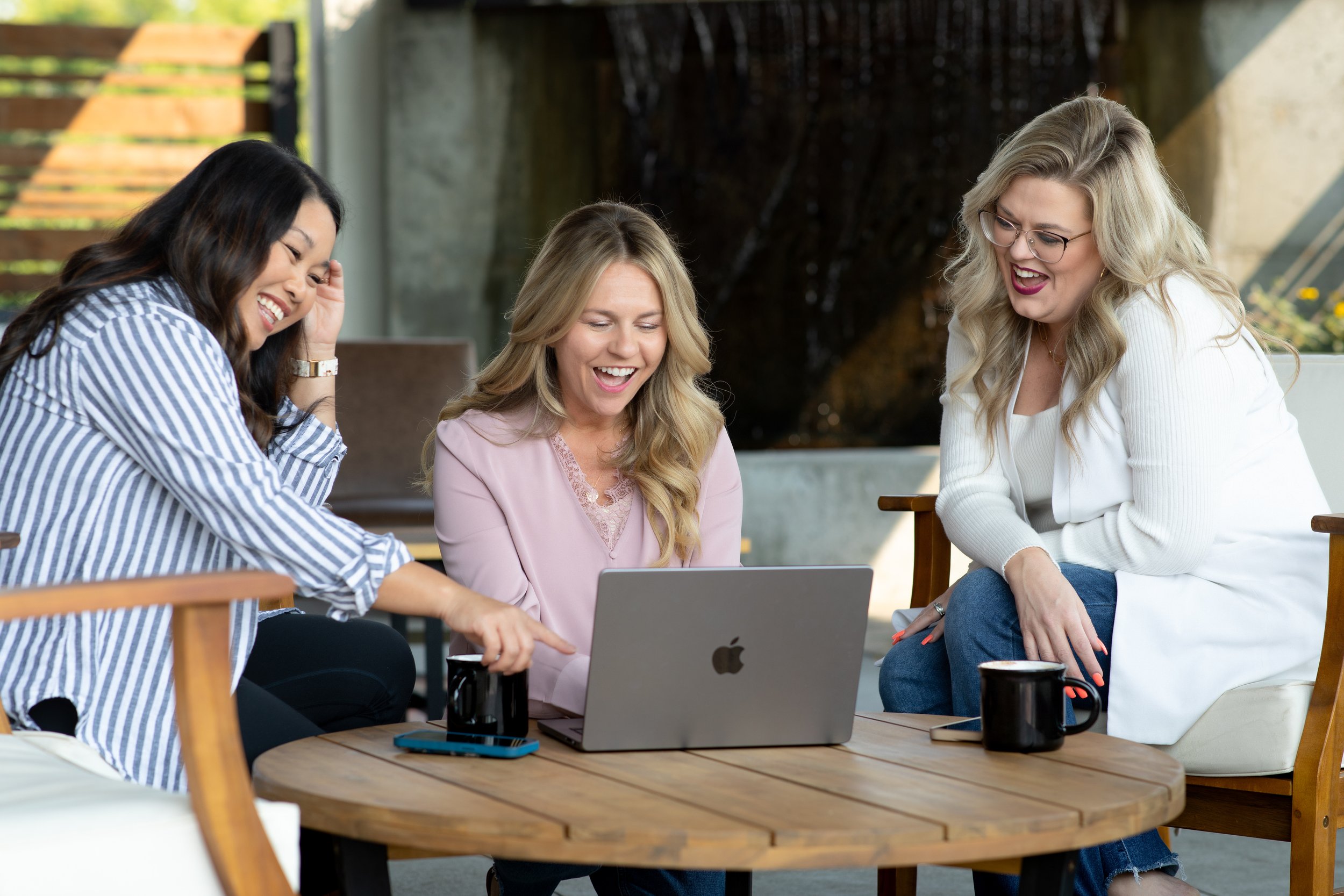 Three women sitting around a wooden table outdoors, laughing and looking at a laptop.