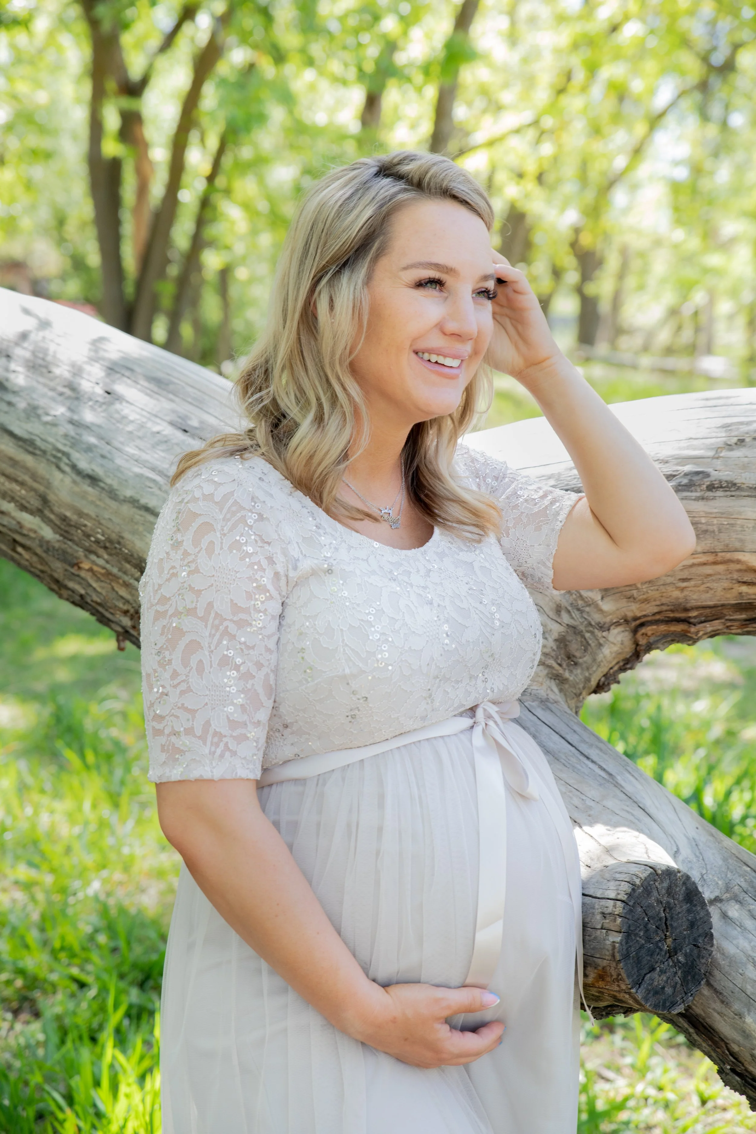 A pregnant woman with blonde hair in a white lace dress, standing outdoors near a fallen tree, smiling and touching her belly in a sunny, green park.