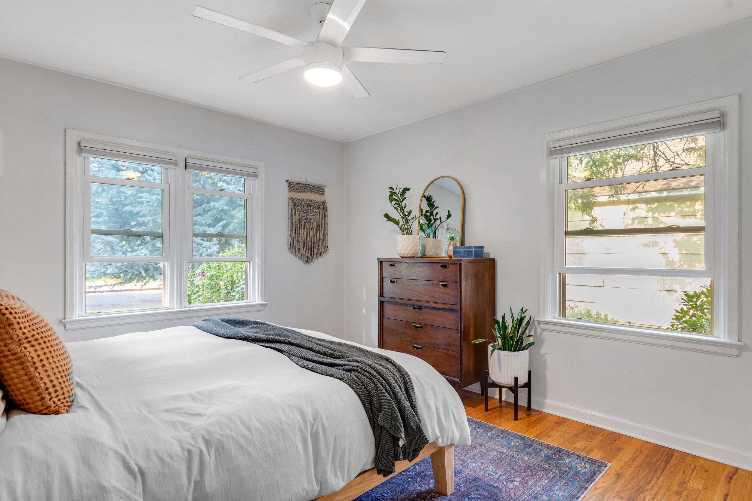 A bright bedroom with white walls, two large windows showing outdoor greenery, a bed with white bedding and a gray throw, a wooden dresser with a mirror and decor, a potted plant on a stand, a wall hanging, and a ceiling fan.