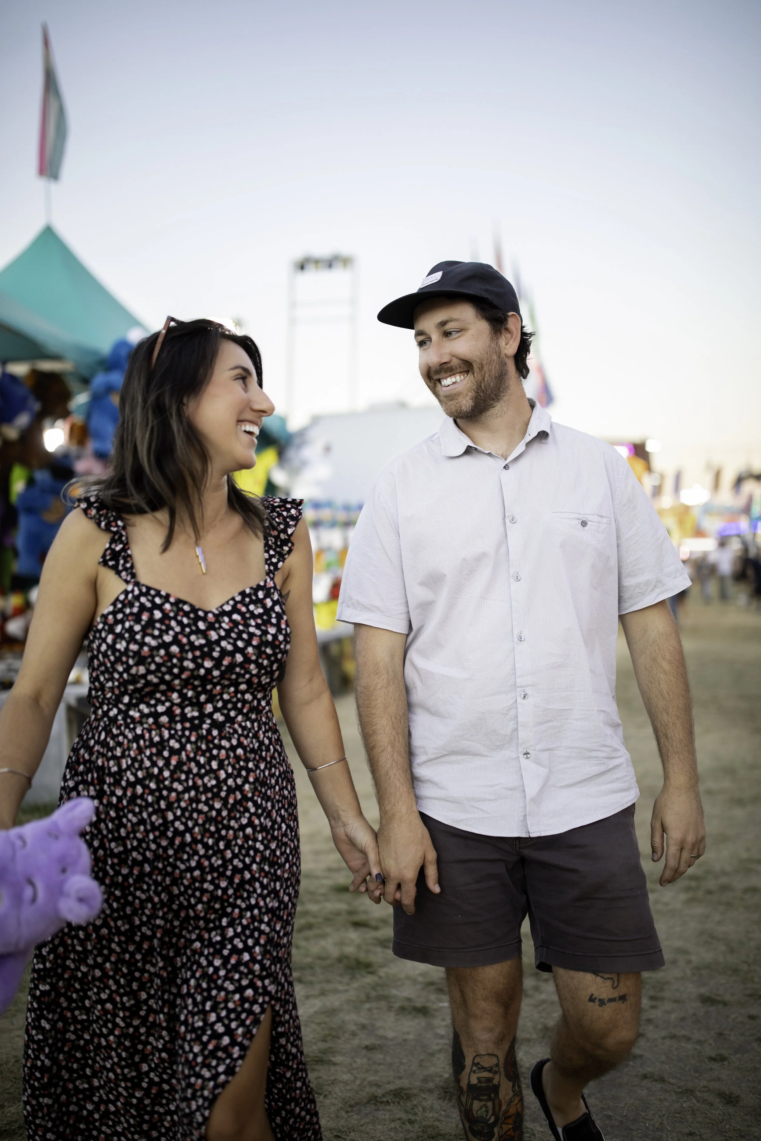 A smiling couple holding hands at an outdoor fair or carnival during the day, with colorful tents and booths in the background.