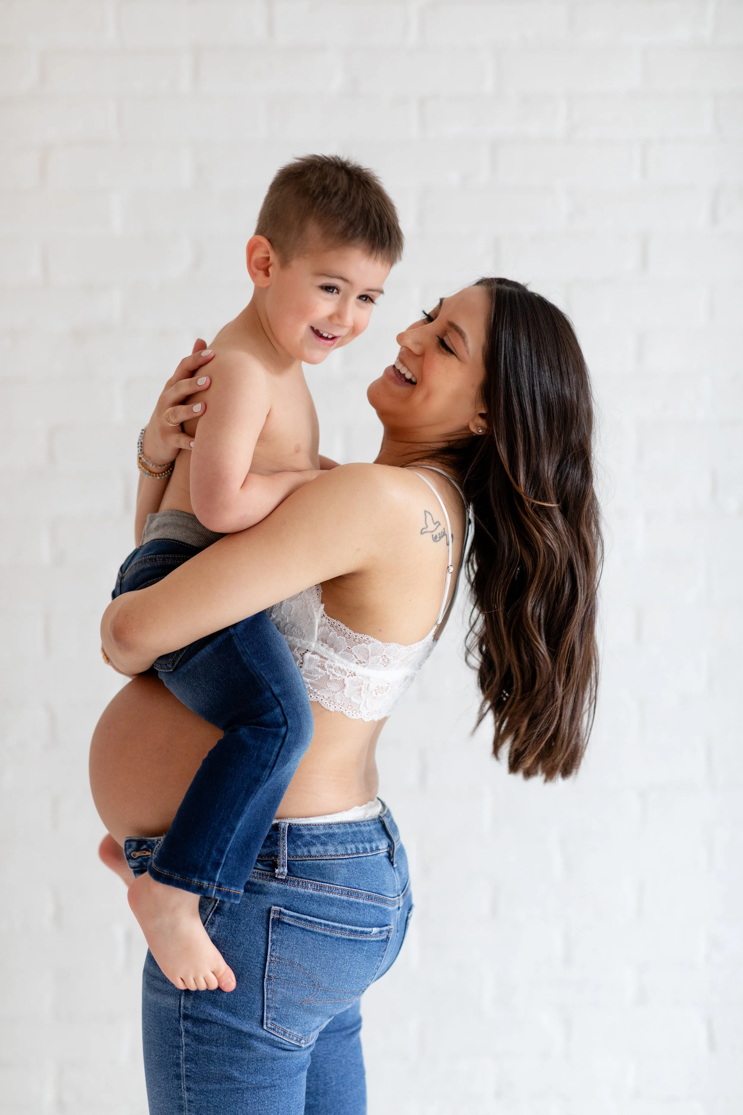 A woman holding a young boy in her arms, both smiling and laughing, against a white brick wall.