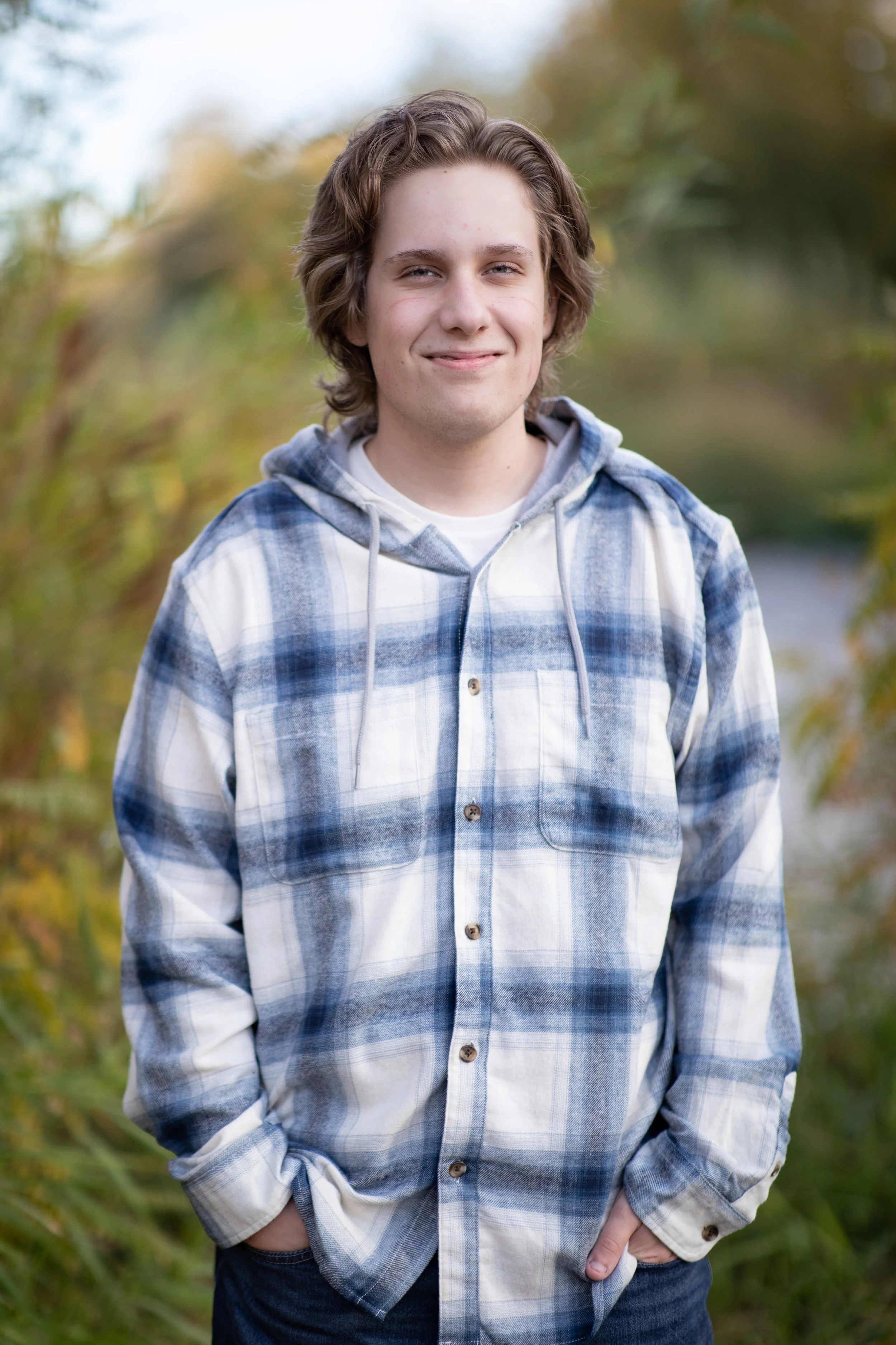 A young man with wavy brown hair smiling outdoors, wearing a blue and white plaid hoodie with a white undershirt, standing in front of greenery with some yellowish leaves.