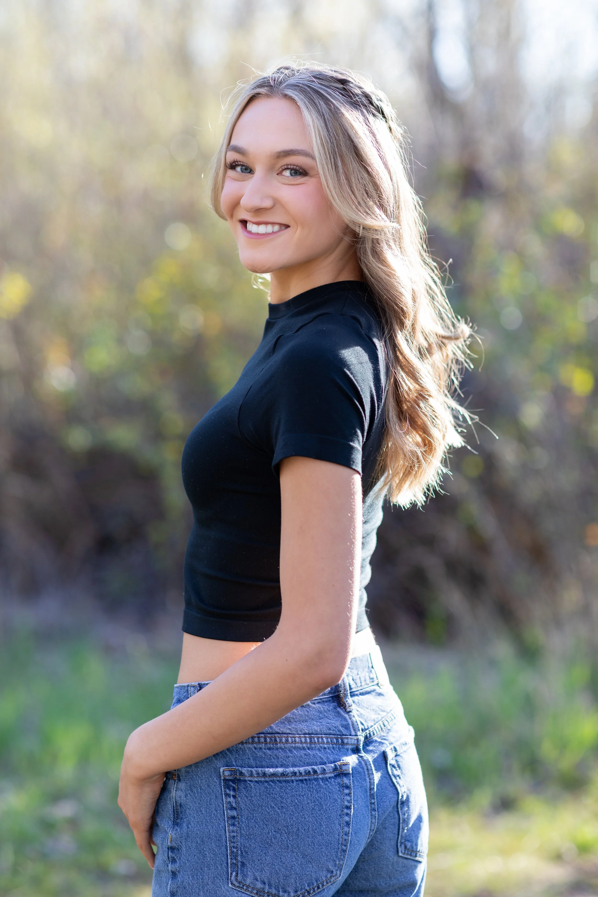 A young woman with blonde wavy hair smiling outdoors, wearing a black crop top and blue jeans, standing in a natural setting with blurred trees in the background.