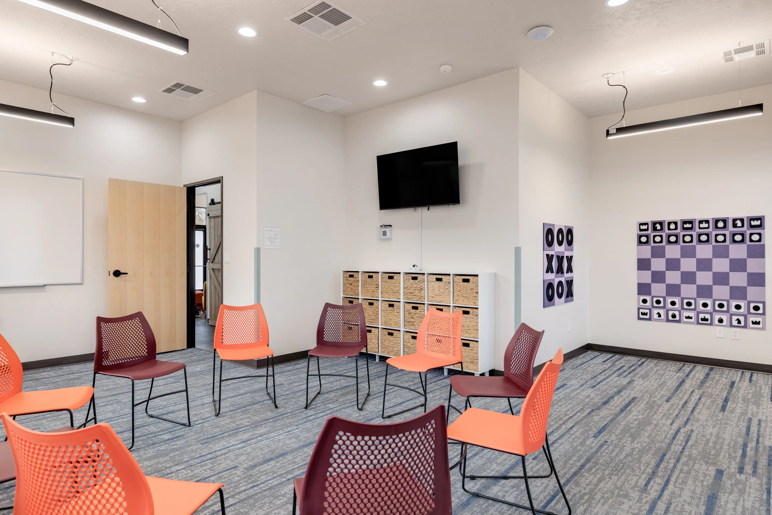 An empty conference or classroom room with colorful chairs arranged in a circle, a wall-mounted TV, storage cubbies, and wall decorations including a chess board and tic-tac-toe game.