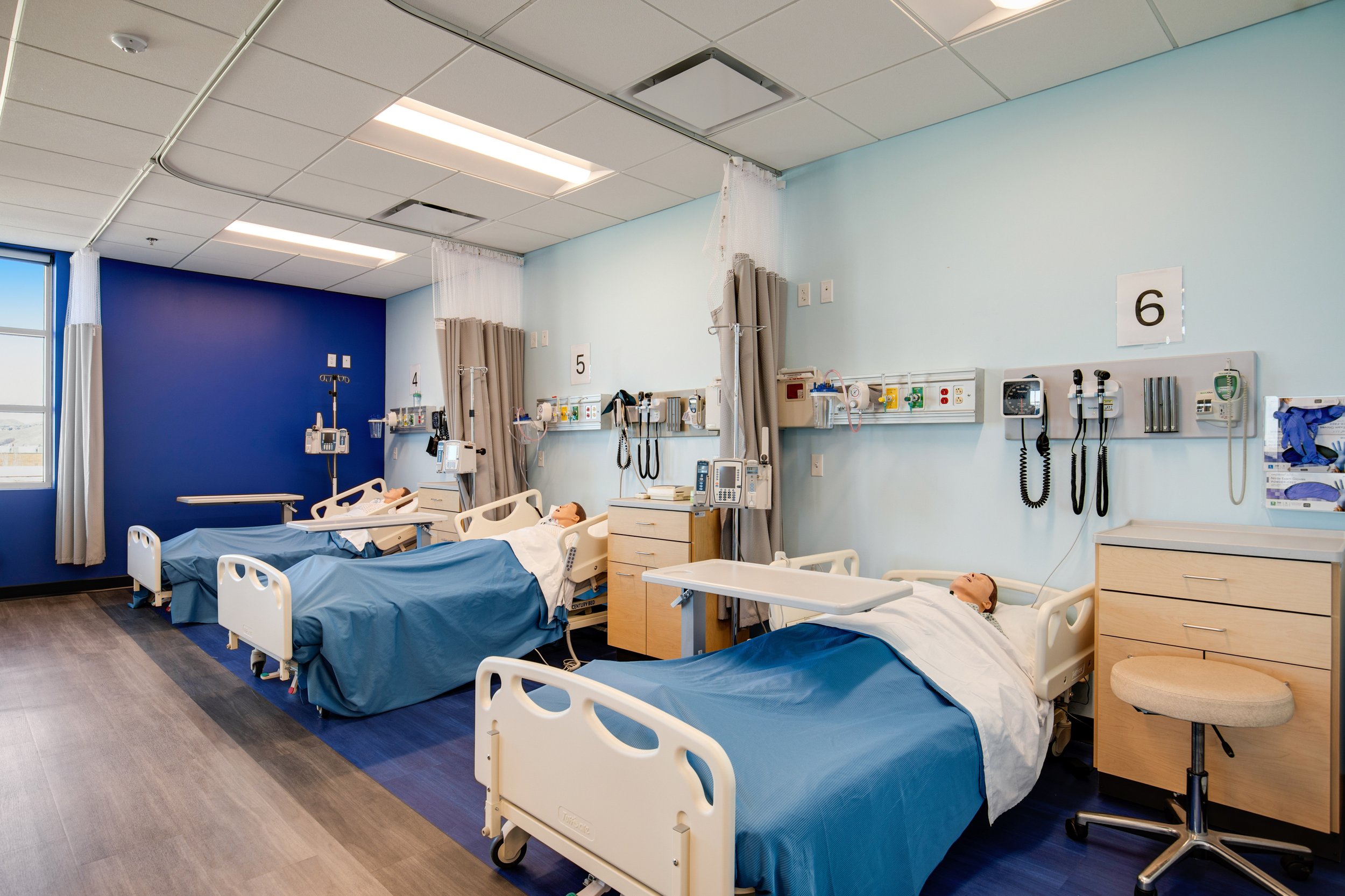 Hospital room with three beds, each with a medical mannequin, medical equipment, and patient monitors, separated by curtains, with a window on the left and labeled sections 4, 5, and 6.