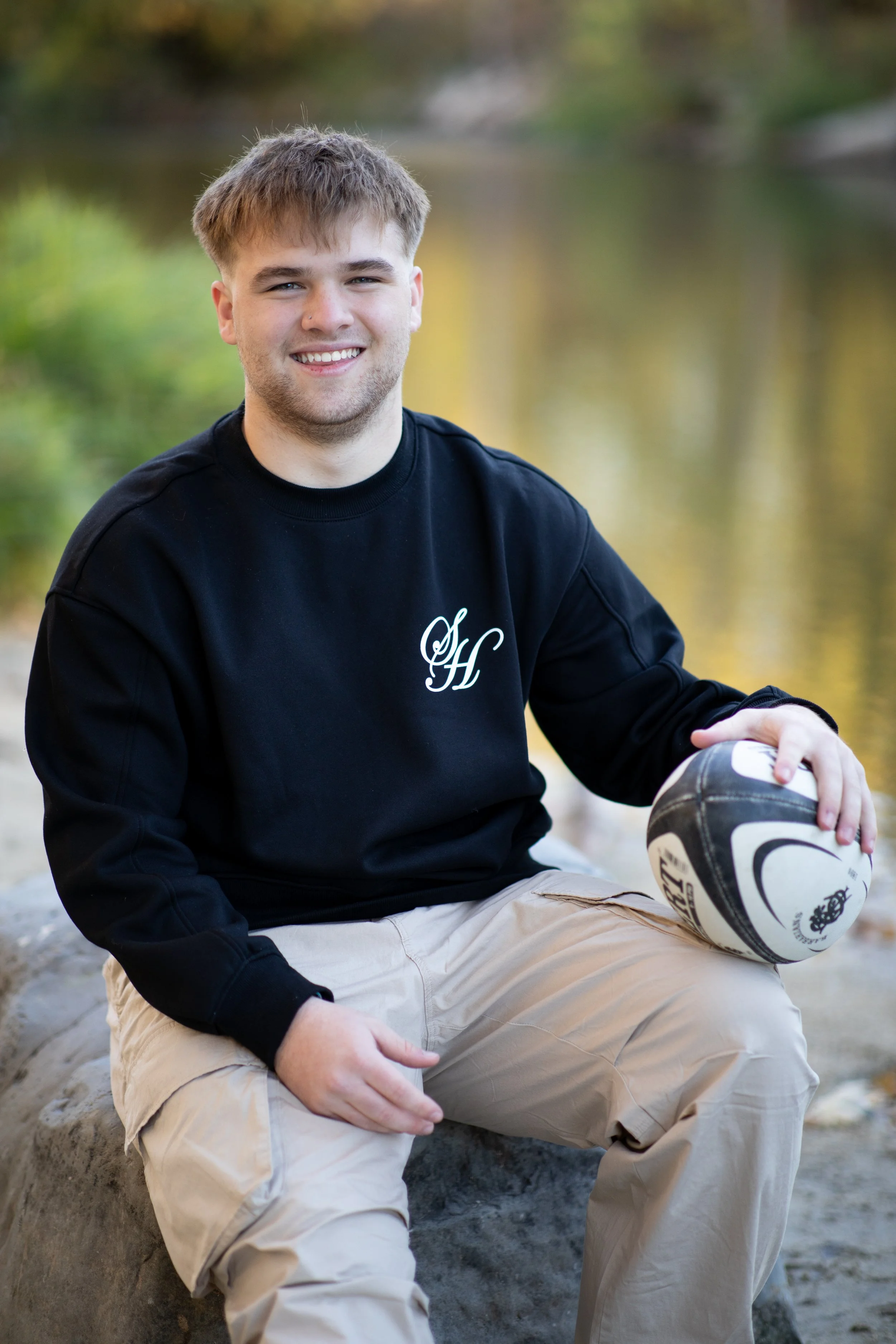 A young man with light brown hair, sitting on a rock near a river, holding a black and white rugby ball, smiling at the camera, wearing a black sweatshirt with initials 'SH' and beige cargo pants.