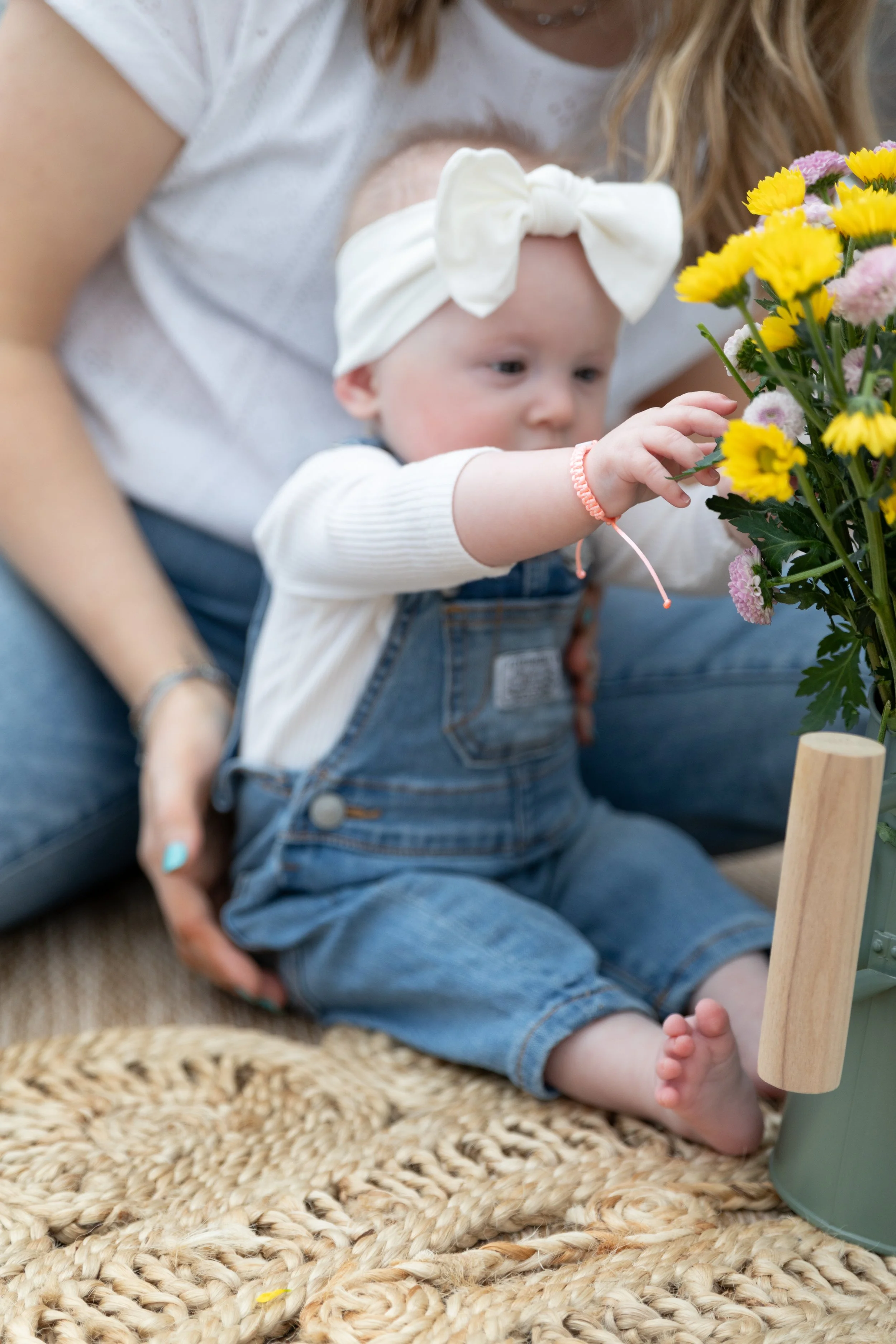 A young child with a white headband, white sweater, and denim overalls reaching out to touch a bouquet of yellow and pink flowers, seated on a woven rug with an adult nearby.