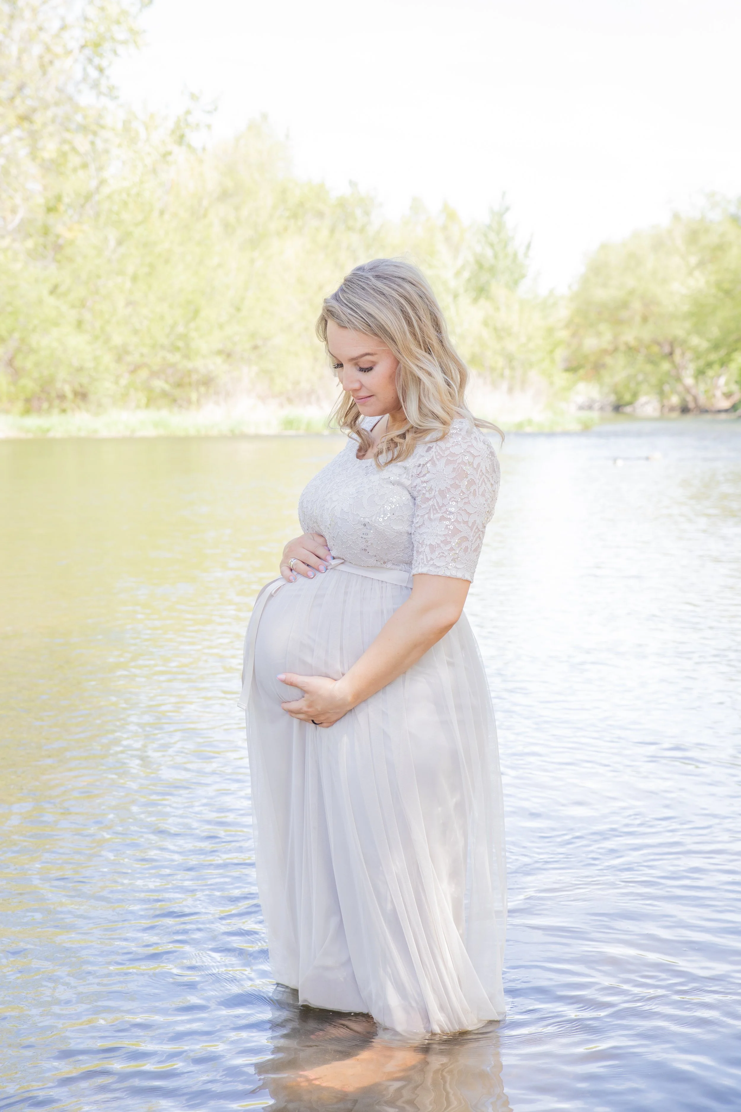 Pregnant woman standing in a lake, wearing a white lace top and pleated skirt, holding her belly with both hands, surrounded by trees and water, during daytime.