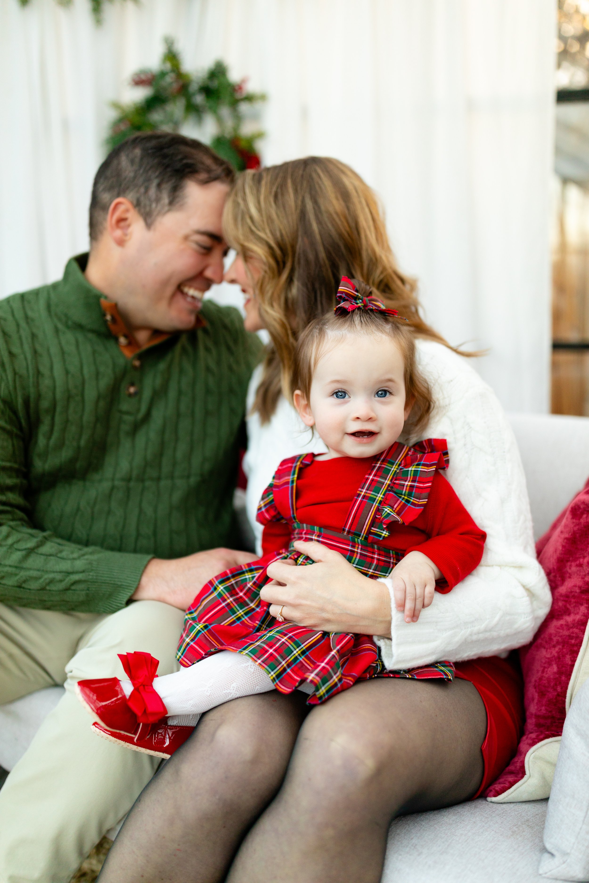 A young girl in red plaid dress sitting on a woman's lap with a smiling couple in the background.
