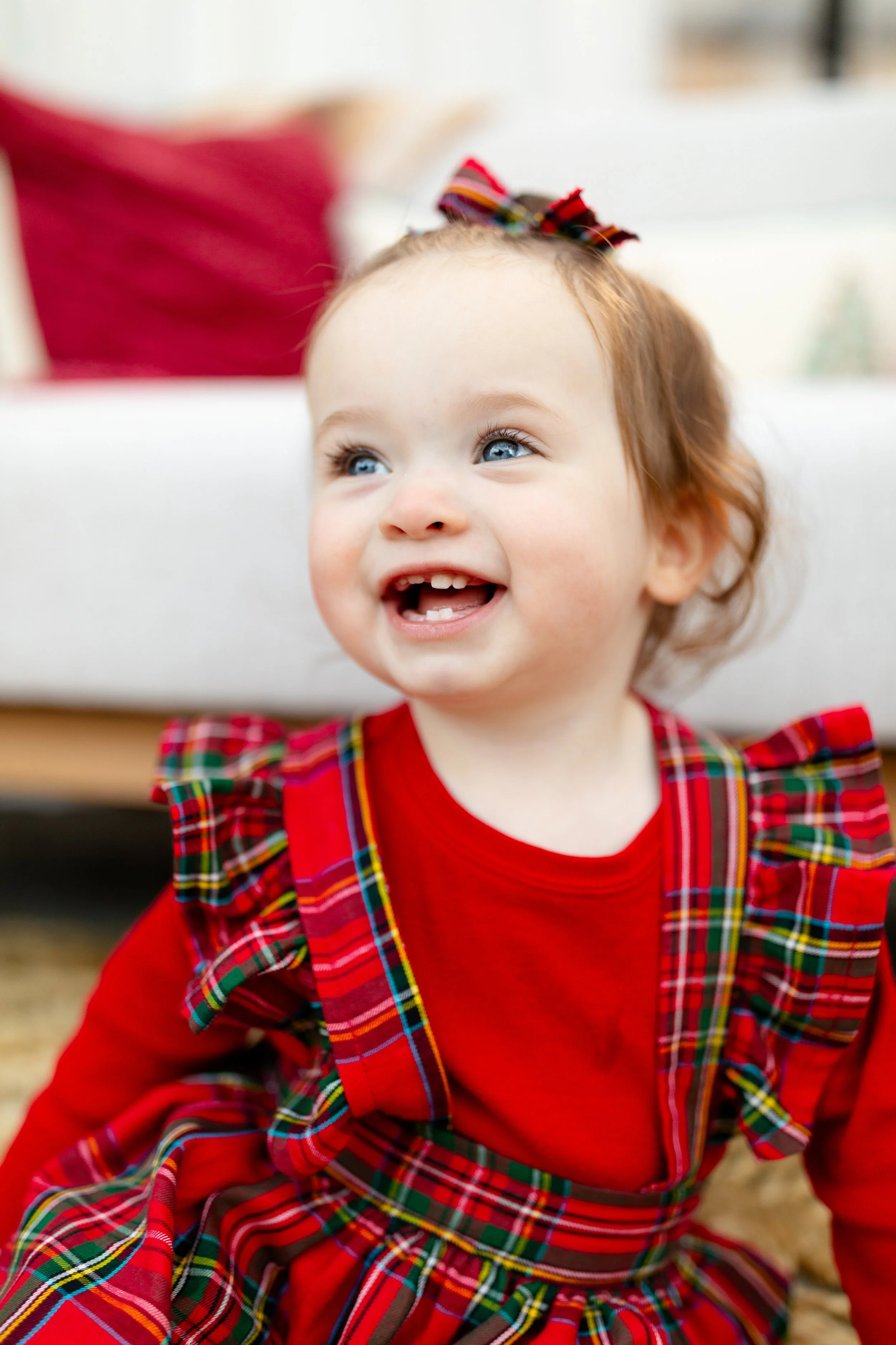 A happy young girl with curly blonde hair, blue eyes, and a big smile showing her teeth, sitting on the ground wearing a red plaid dress with ruffled shoulders and a bow in her hair.