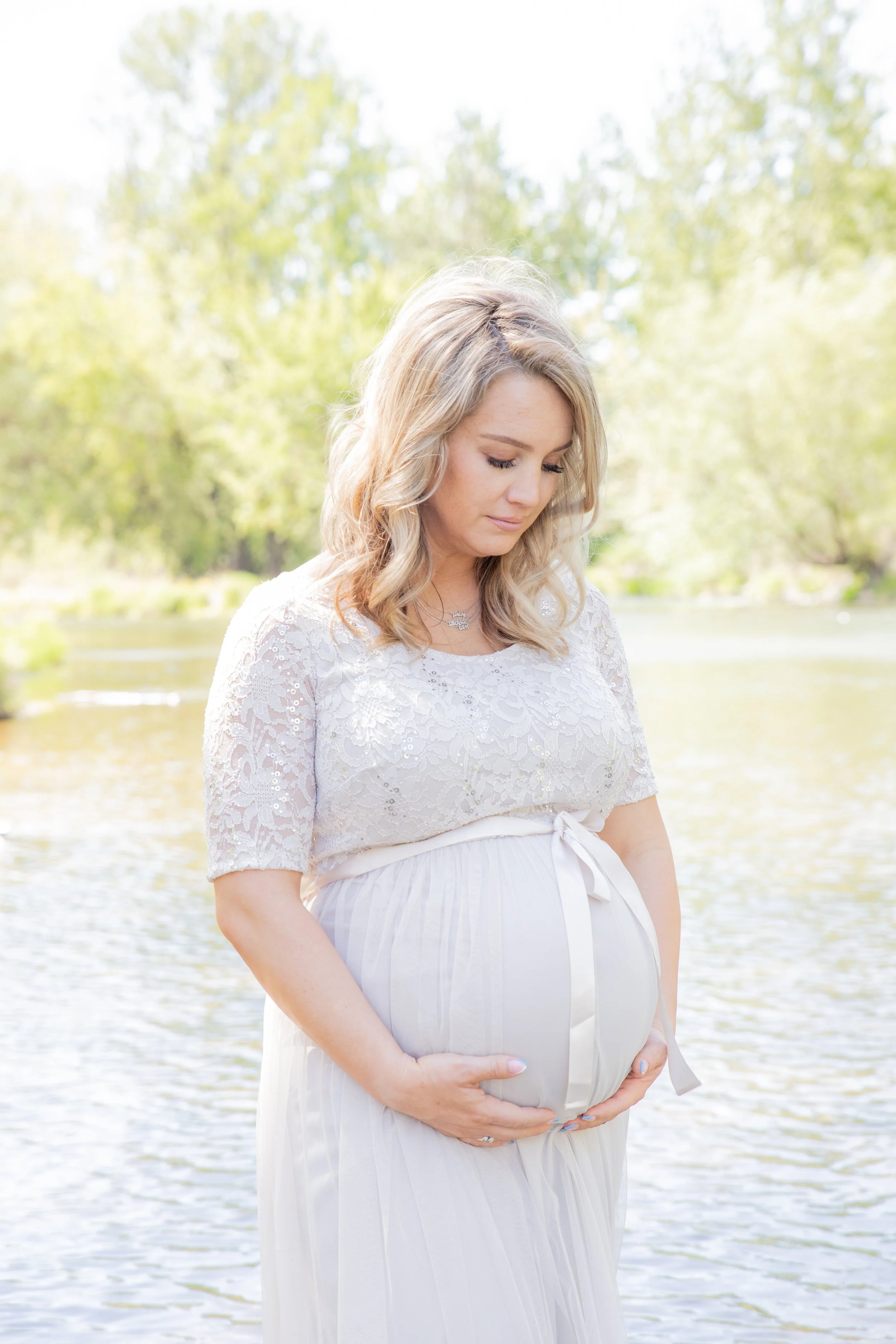 A pregnant woman with blonde hair, wearing a lace white dress, gently holding her belly, standing outdoors near a water body with green trees in the background.